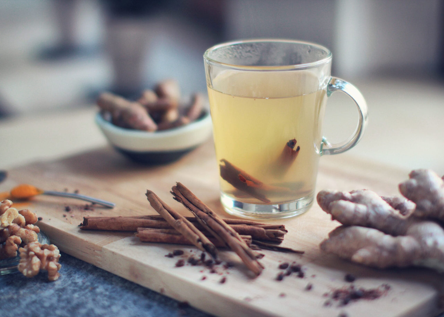 Glass mug of ginger tea on a cutting board with fresh ginger, cinnamon sticks, walnuts, and a bowl of spices