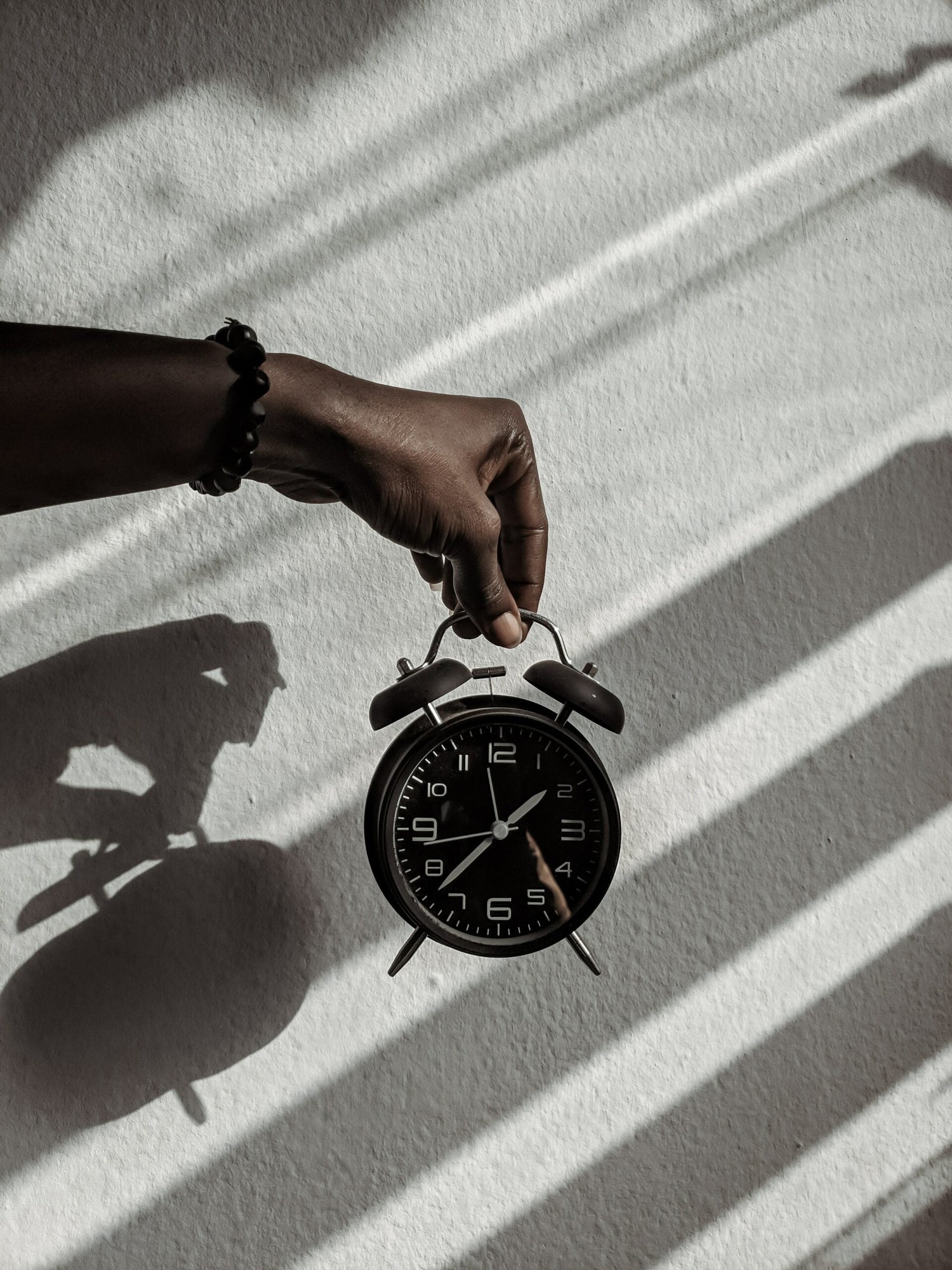 Hand holding a black vintage alarm clock casting striped shadows on a textured white wall