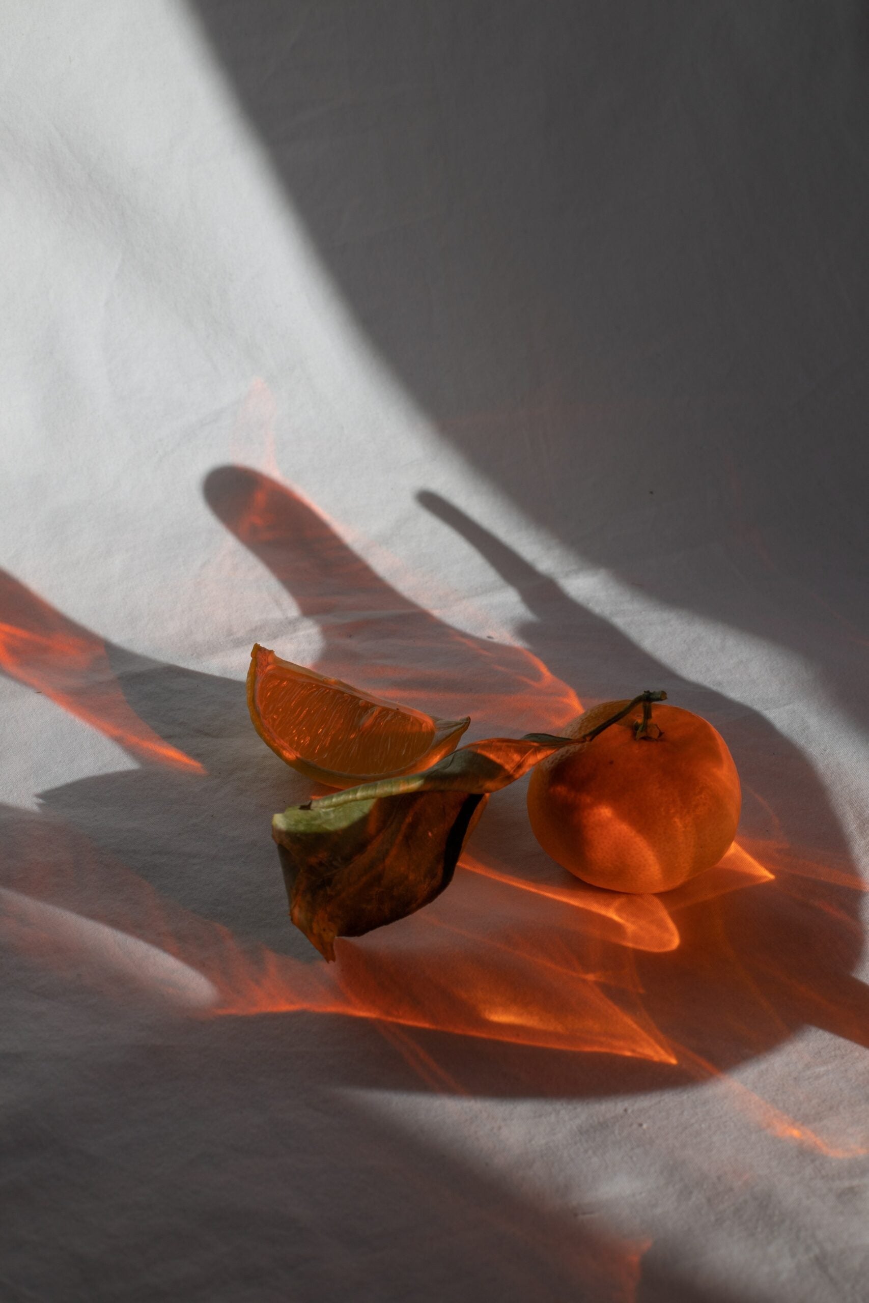 Two tangerines, one whole with a leaf and one sliced, resting on white fabric with warm orange light and dramatic shadows