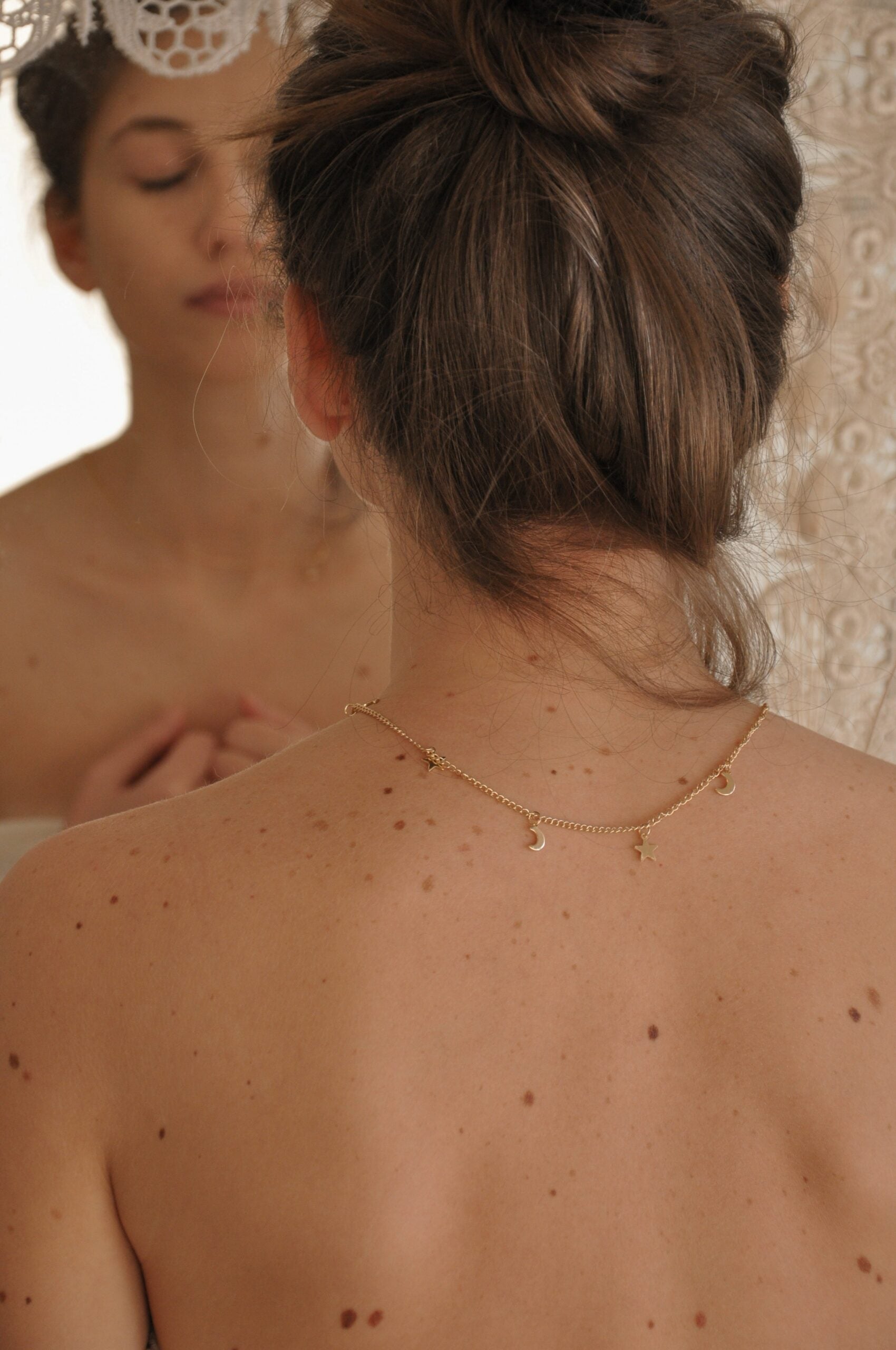 Woman with hair in a bun facing a mirror, bare freckled back wearing a delicate gold moon-and-star charm necklace