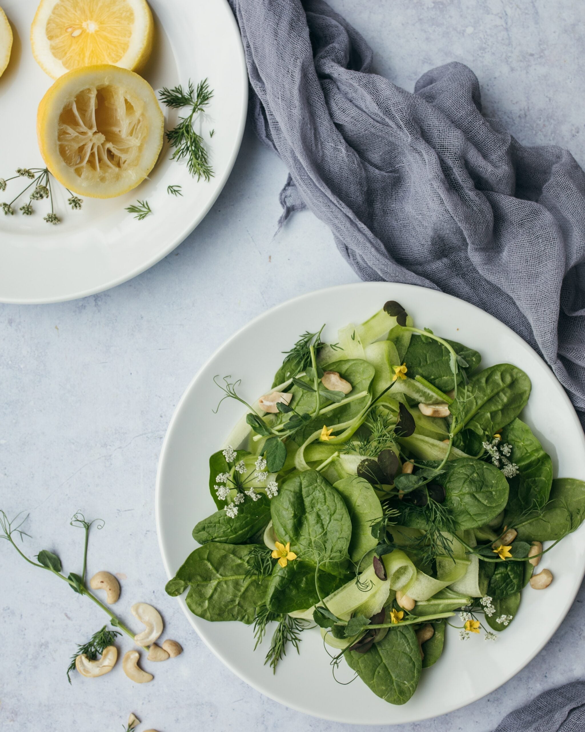 Top-down view of a green spinach salad with cucumber ribbons, herbs and cashews on a white plate beside a squeezed lemon and gray cloth
