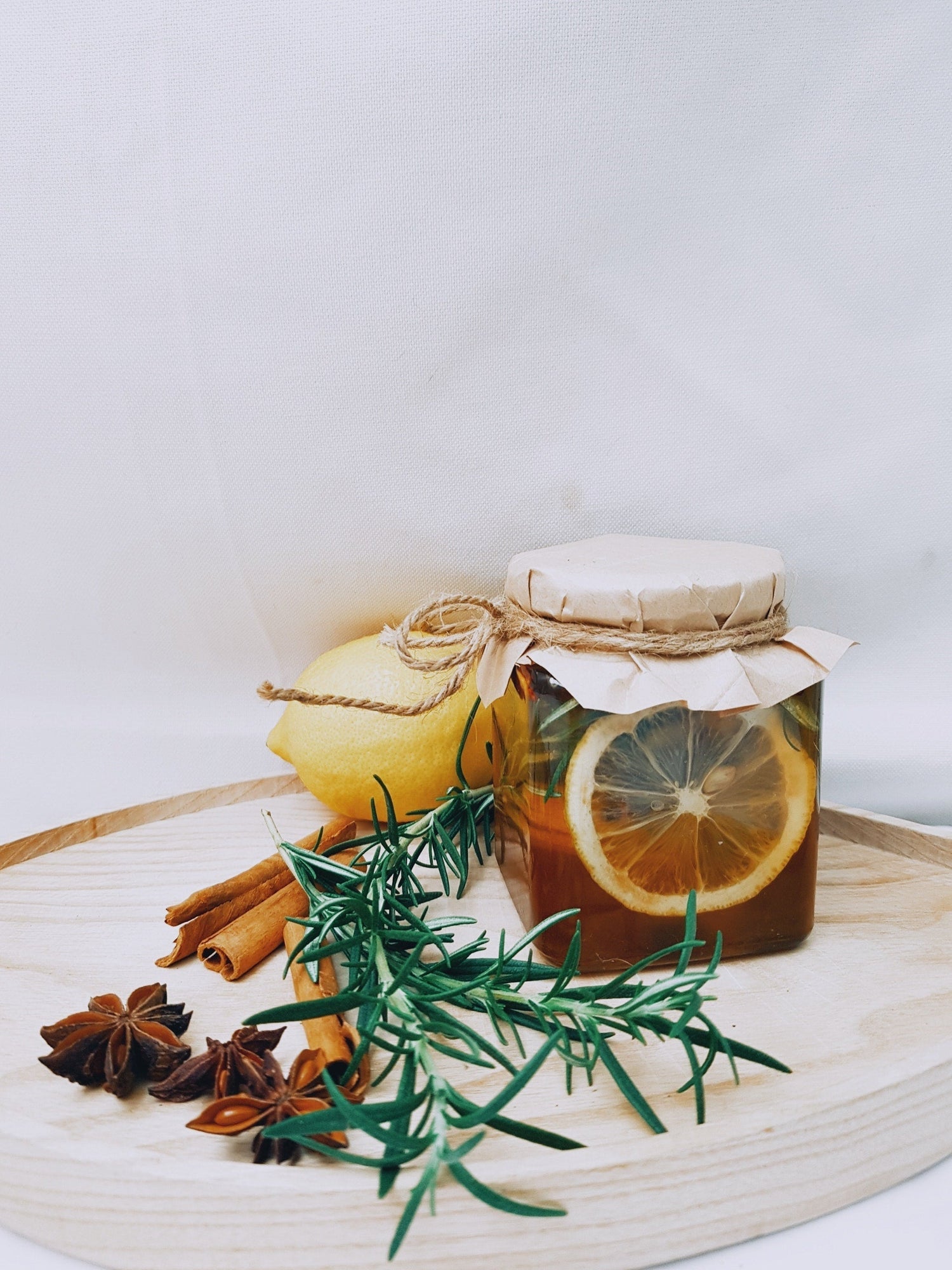 Jar of lemon-infused honey with rosemary, cinnamon sticks, and star anise on a round wooden board against a white background
