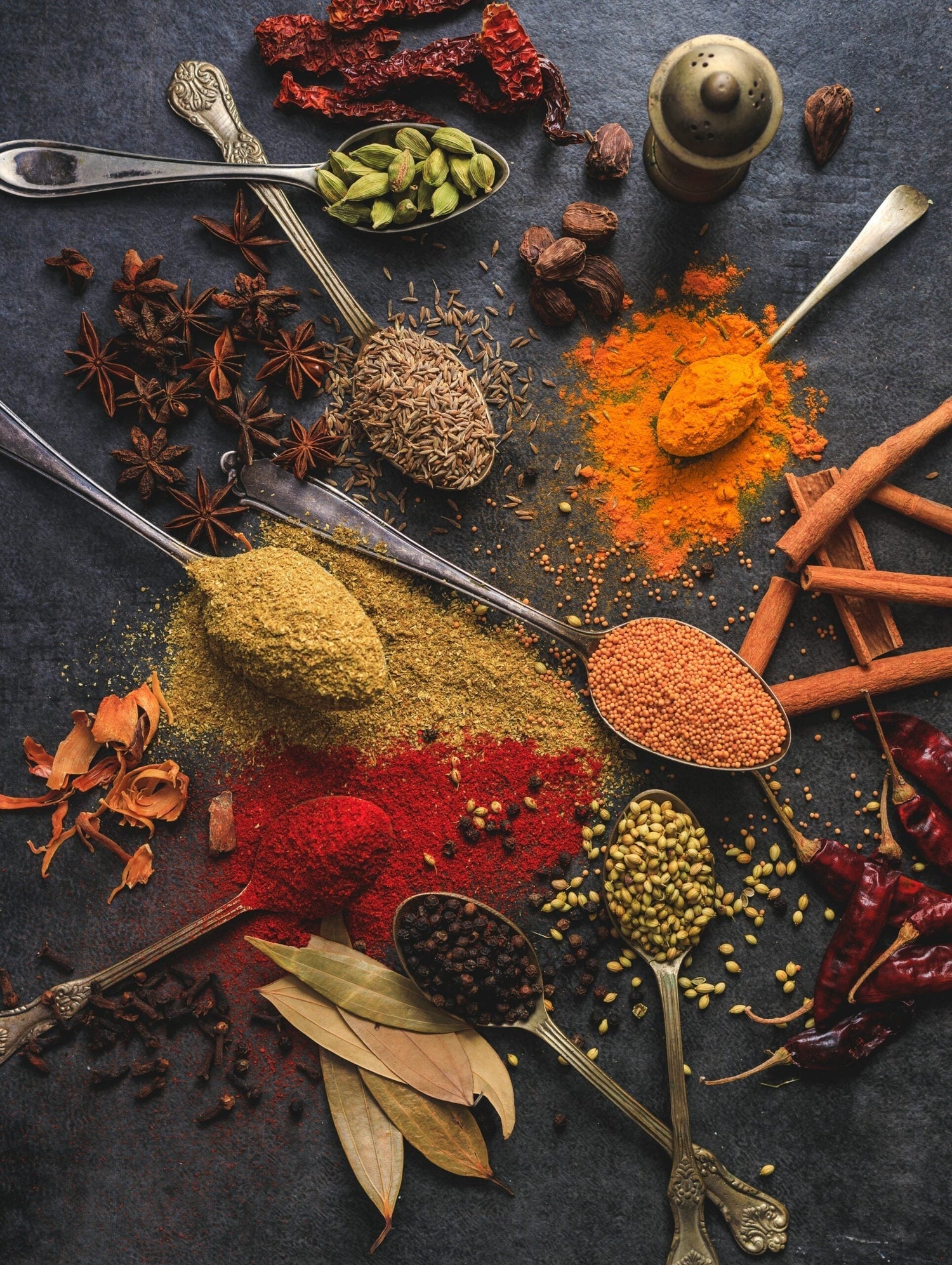 Assorted whole and ground spices in spoons on a dark surface, including turmeric, paprika, mustard seeds, cardamom
