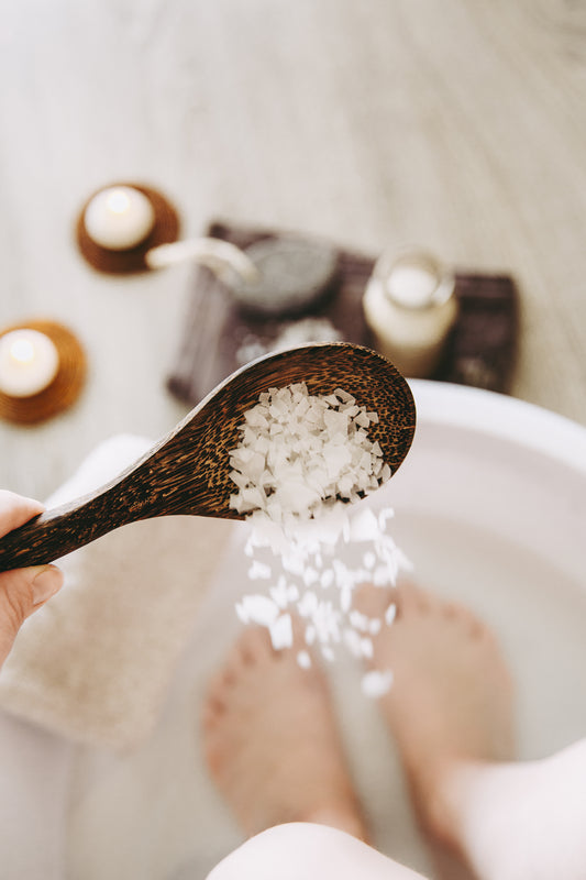 Feet soaking in a foot bath as a hand sprinkles coarse bath salts from a wooden spoon, candles and towel in the background