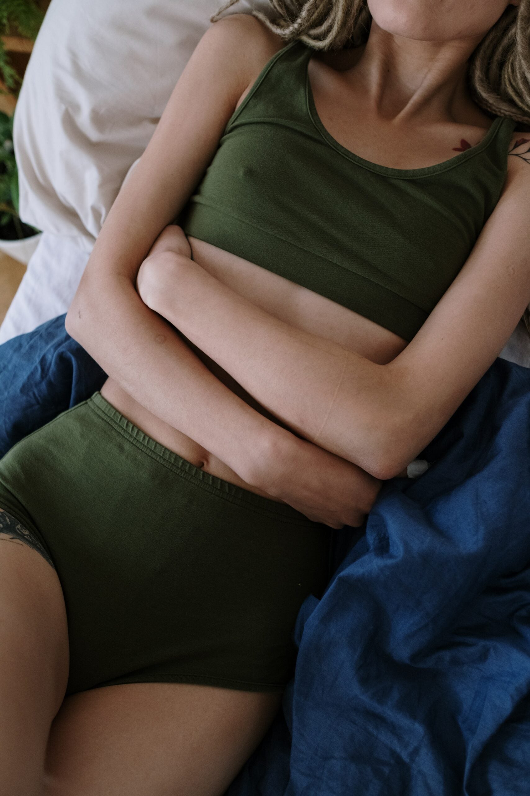 Woman lying on a bed holding her lower abdomen, wearing olive green tank and underwear, appearing to have abdominal pain