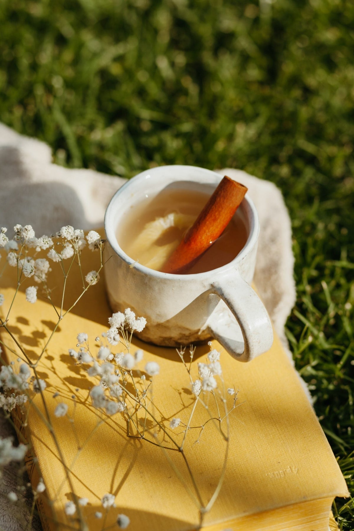A cinnamon stick rests in a mug of warm drink on a book outdoors with baby’s breath flowers