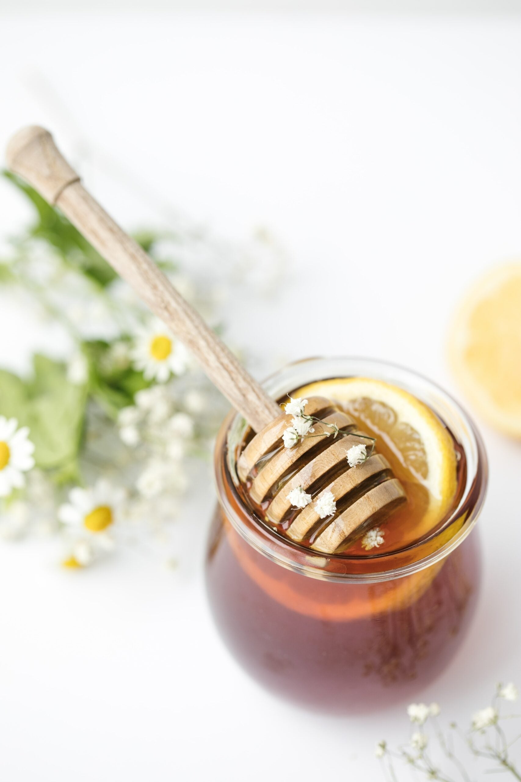 Jar of honey with wooden honey dipper and lemon slice on a white background with chamomile flowers