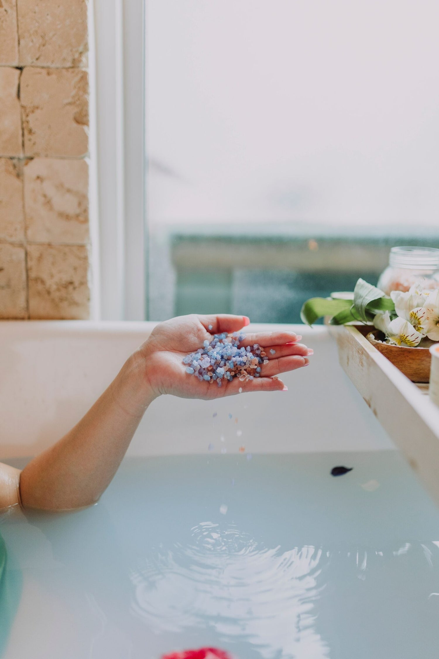Hand sprinkling purple bath salts into a milky bathtub, flowers on a wooden bath tray by a window