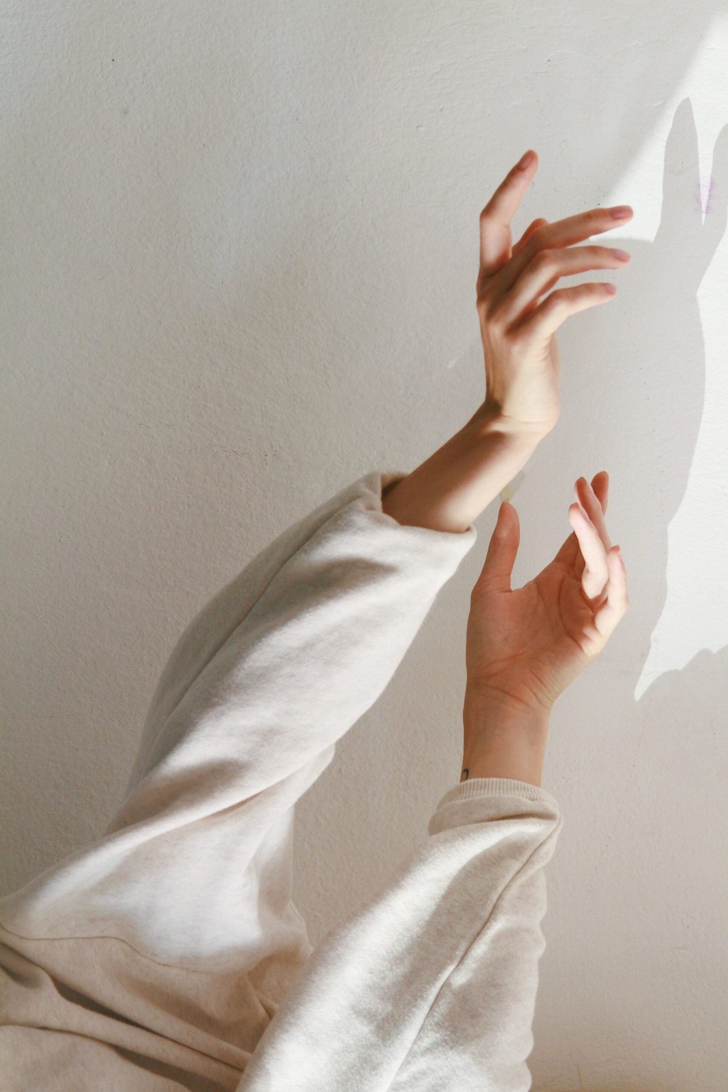 Two raised hands reaching toward sunlight, showing fingernails and casting shadows on a textured white wall