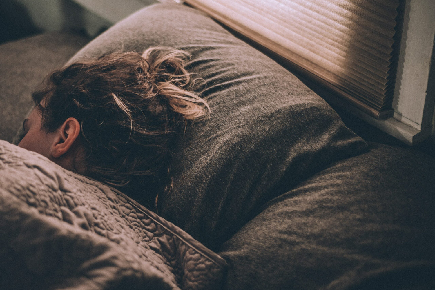 Person sleeping on their side under a quilt in a dim bedroom next to a window with closed blinds