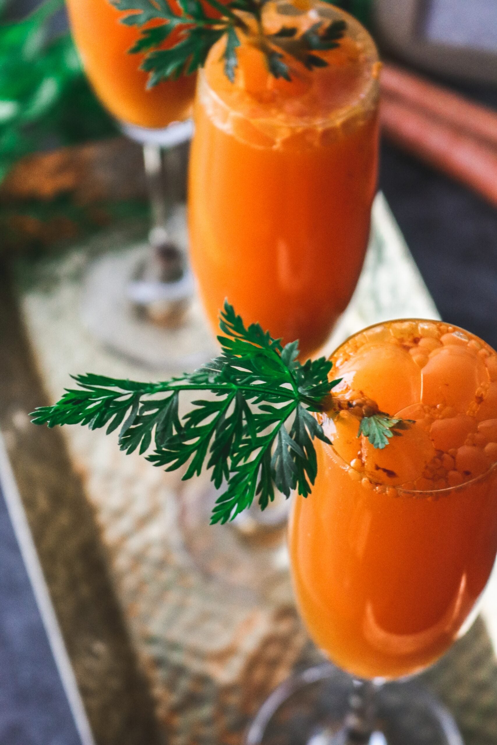 Close-up of two glasses of bright orange carrot juice garnished with fresh carrot greens on a hammered metal serving tray