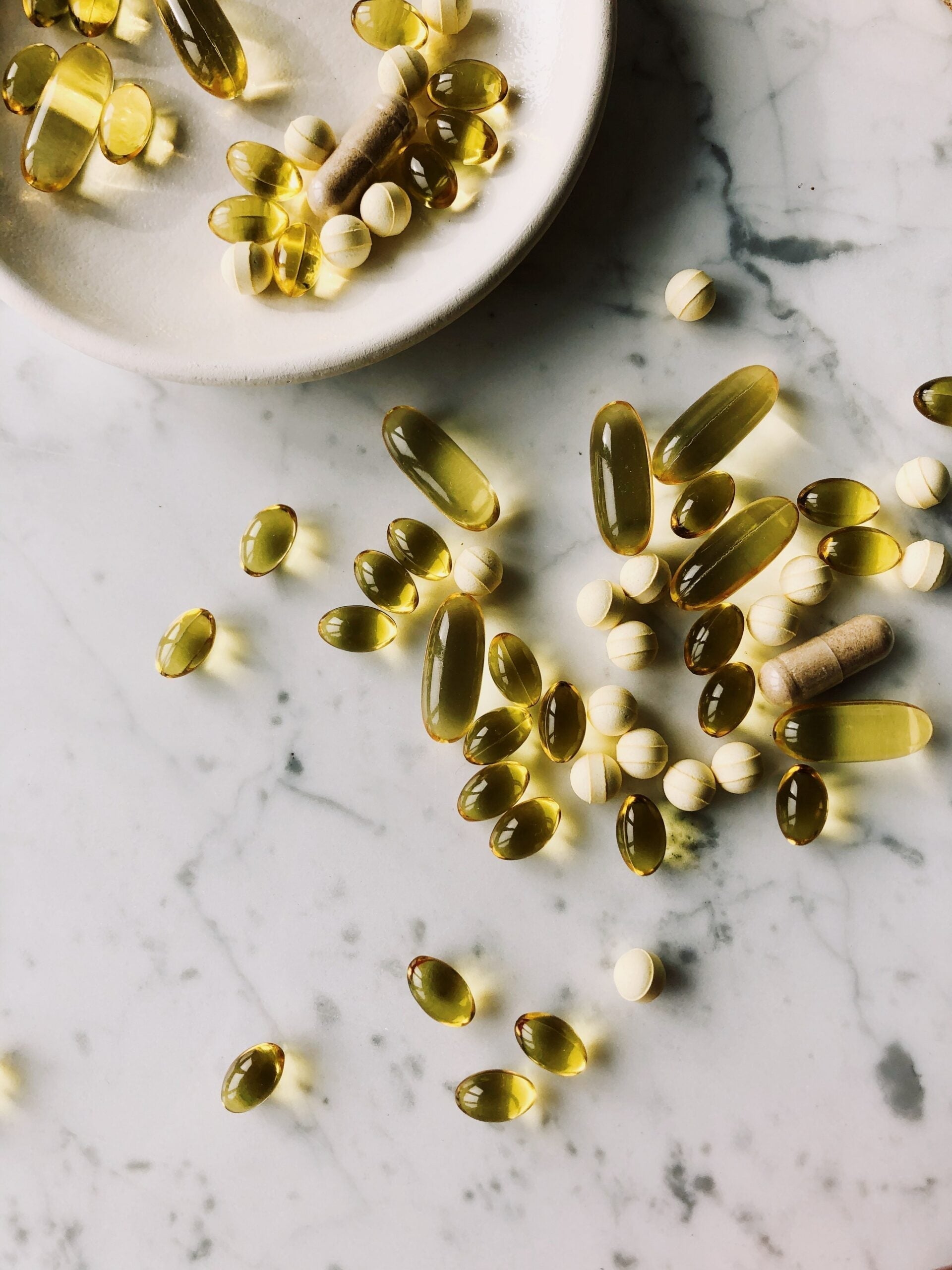 Assorted softgel capsules and round tablets scattered on a white marble countertop with a ceramic dish at top left