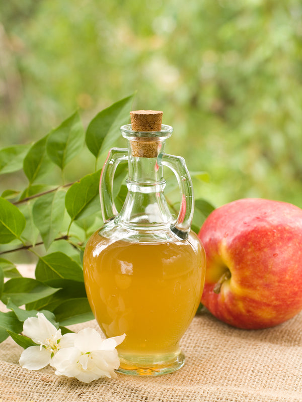 Bottle of apple cider vinegar with a red apple and white blossoms on burlap, green leafy background