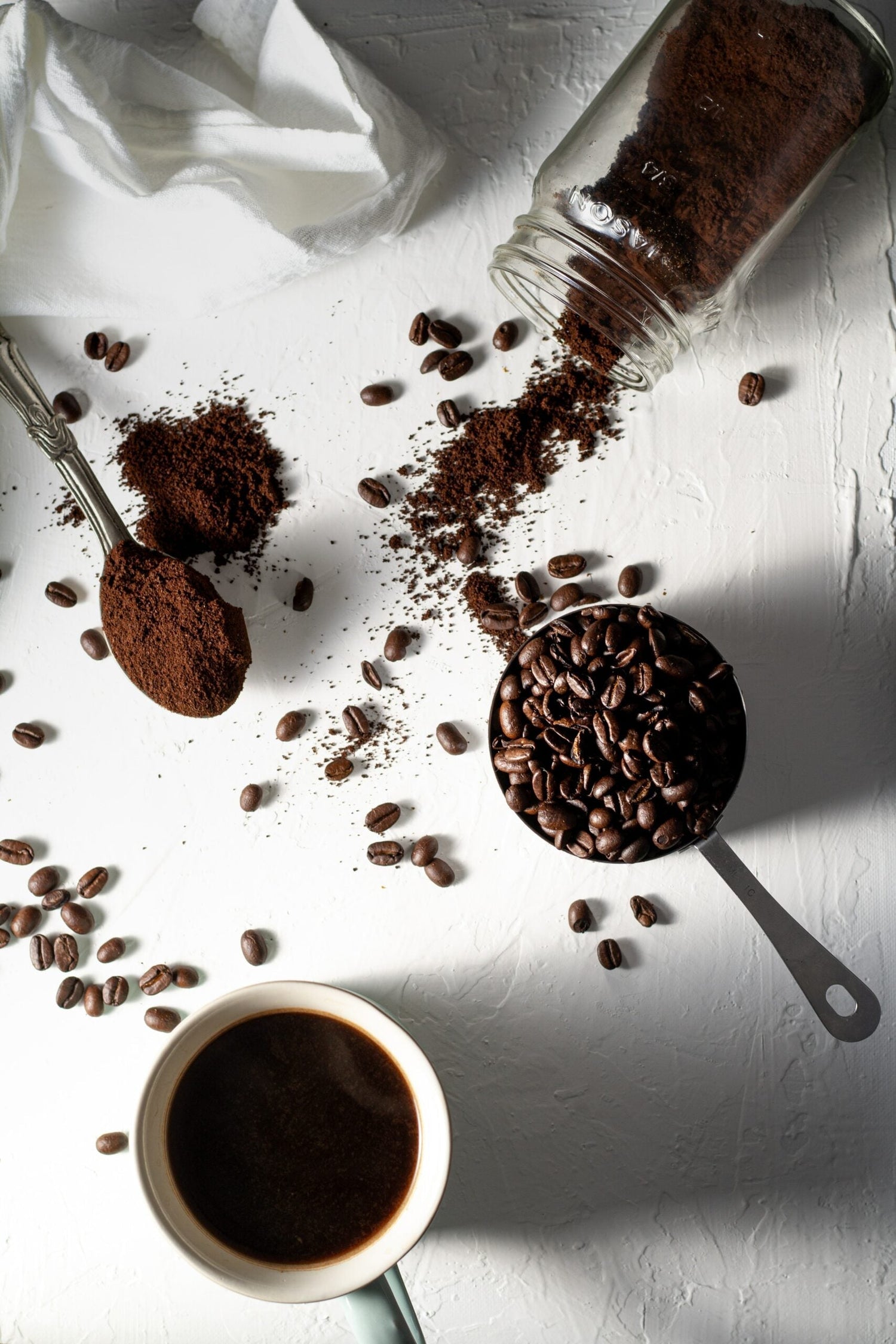 Overhead of a coffee cup, scoop of roasted beans, spilled grounds from a glass jar and spoon on a white surface