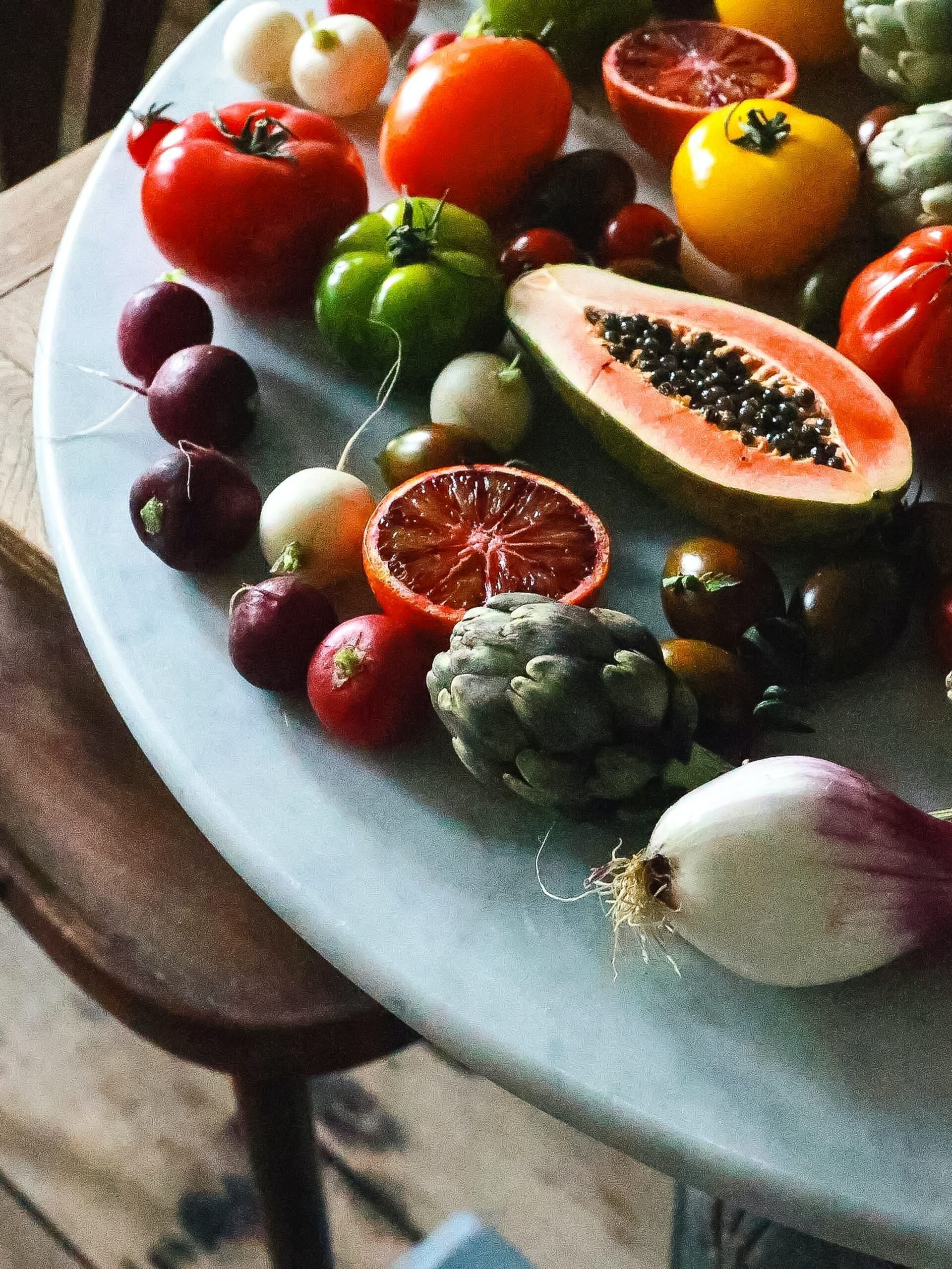 Close-up of assorted raw fruits and vegetables arranged on a marble tabletop with a rustic wooden chair in soft natural light