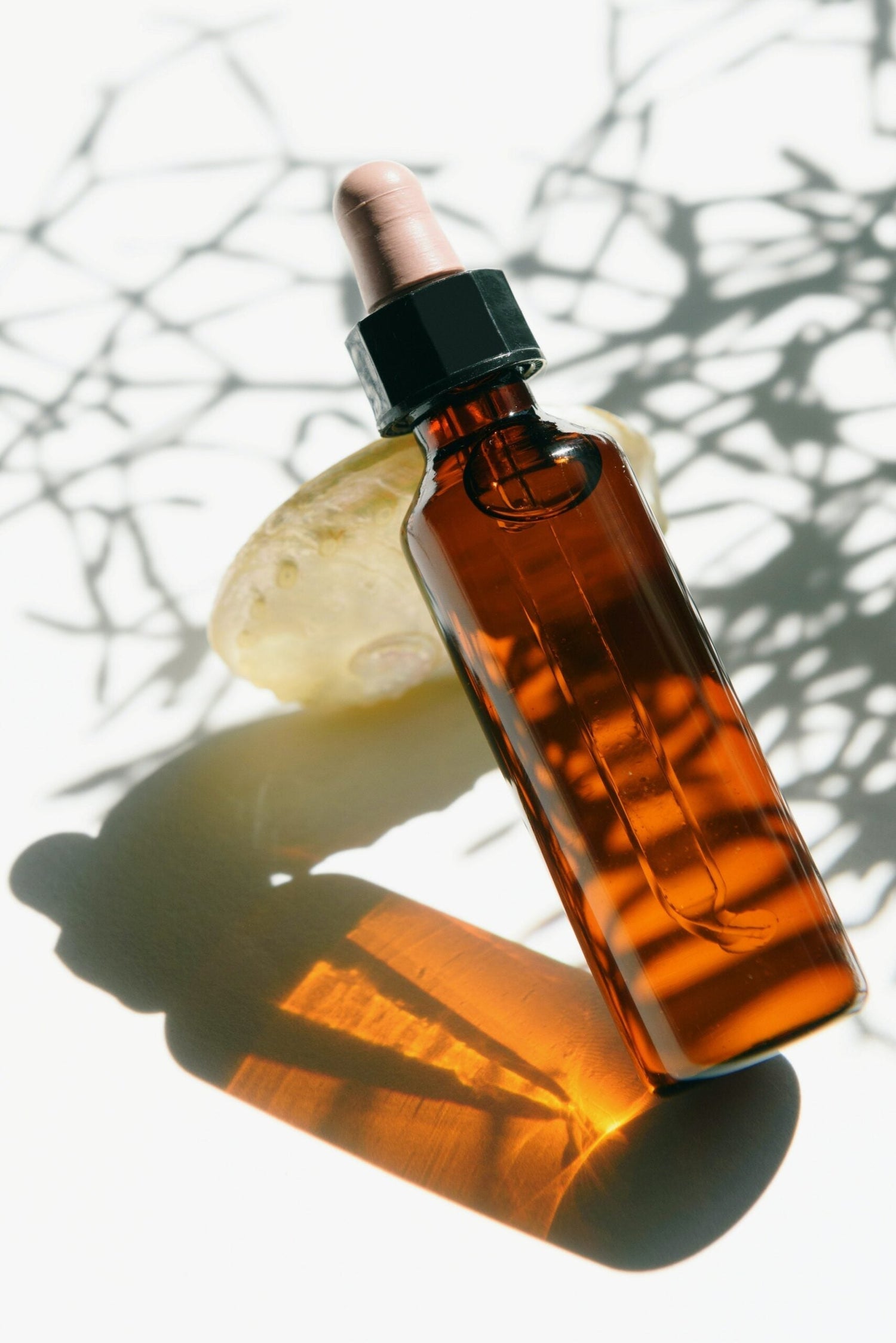 Angled view of amber glass dropper bottle of castor oil showing pipette and warm amber reflection on white background with plant shadow