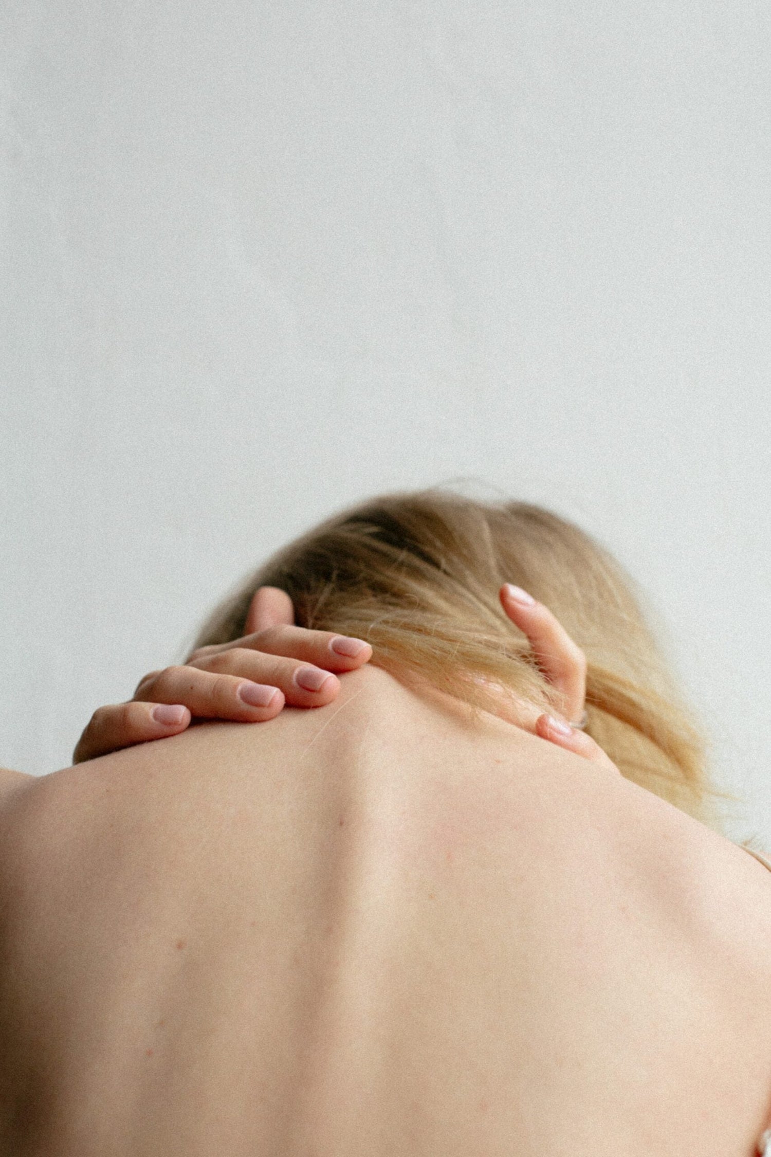 Close up of a person’s upper back and neck with hands resting at the base of the skull against a plain background