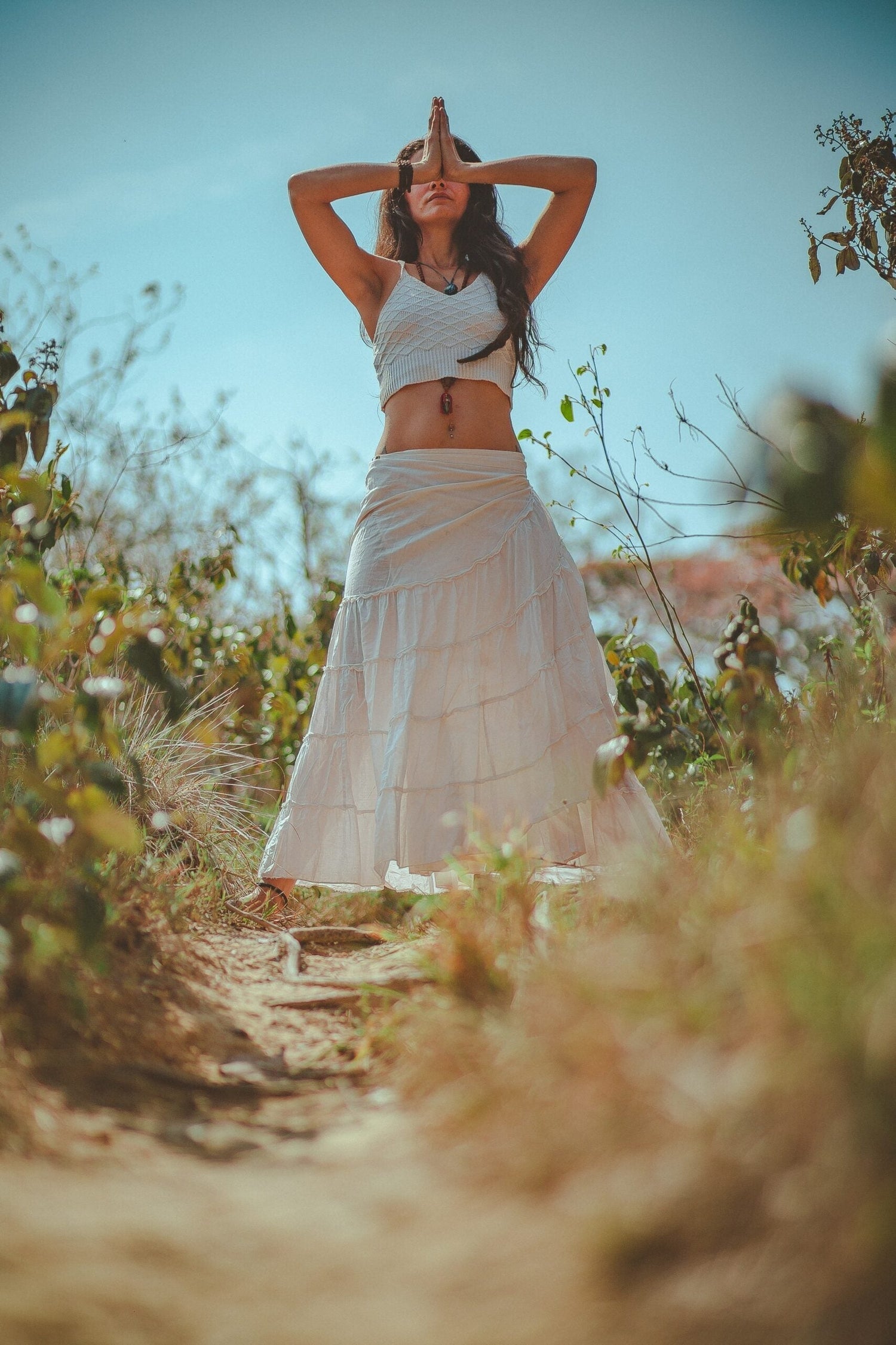 Woman meditating on a sunny path, standing with hands pressed in prayer over her face among dry grasses and shrubs