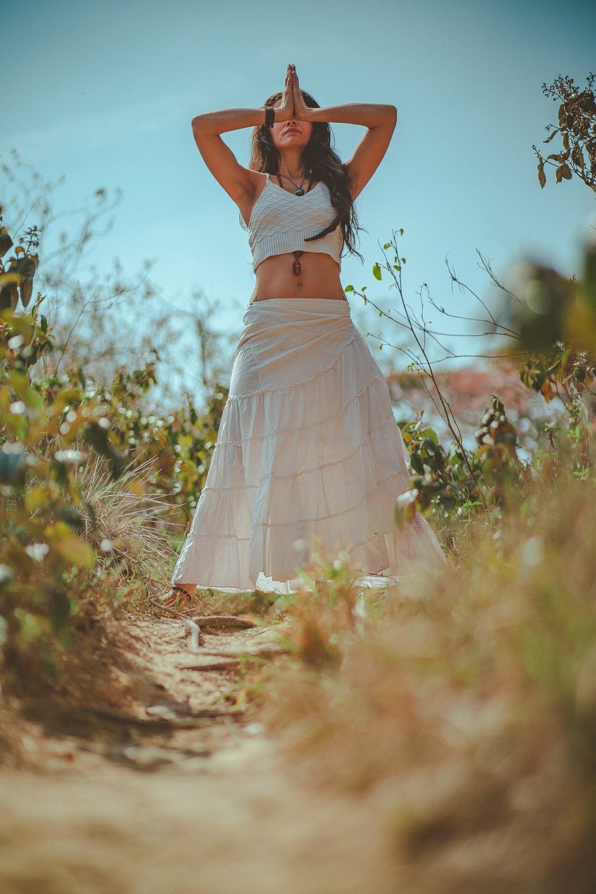 Woman meditating on a sunny path, standing with hands pressed in prayer over her face among dry grasses and shrubs