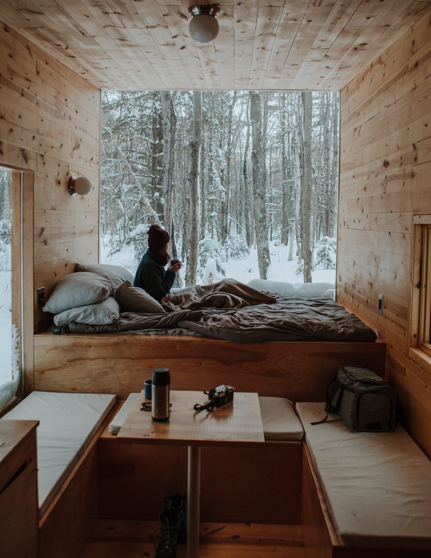 Person sitting on a bed in a wood-paneled cabin, holding a mug and looking out at a snowy forest through a large window