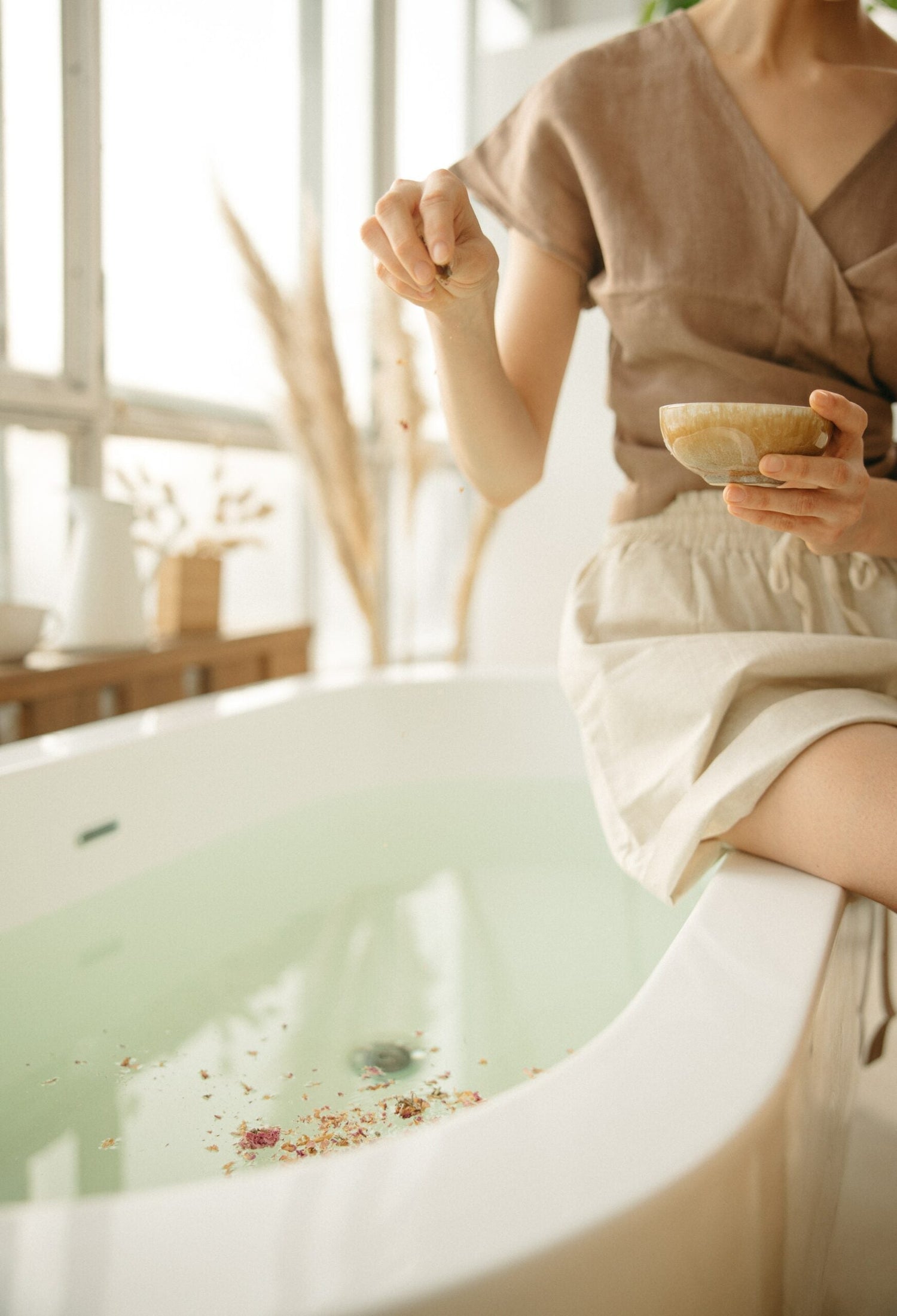 Woman sitting on a bathtub edge sprinkling dried botanicals into pale green bathwater in a sunlit bathroom