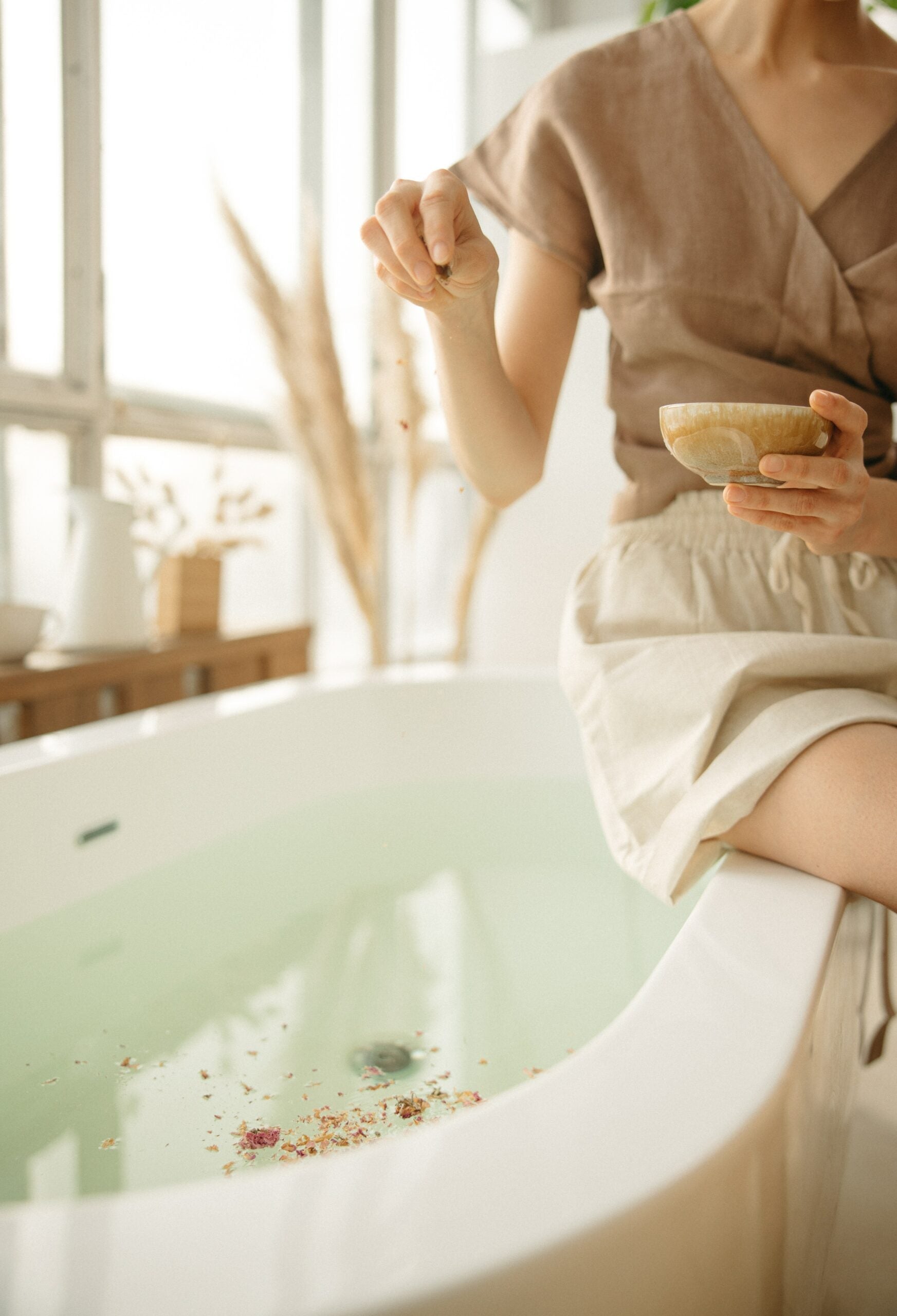 Woman sitting on a bathtub edge sprinkling dried botanicals into pale green bathwater in a sunlit bathroom