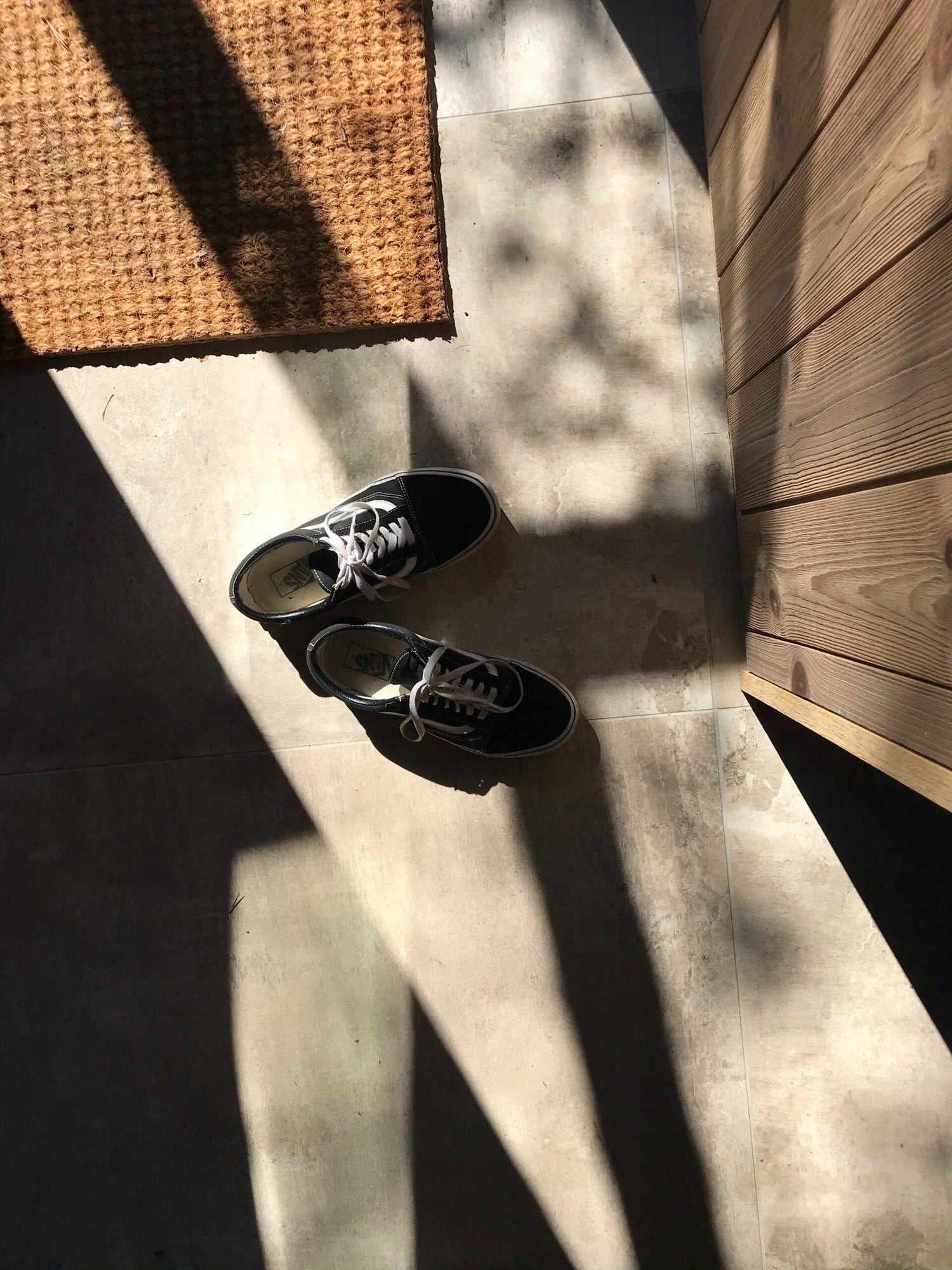 Black canvas sneakers on a sunlit tiled entryway floor beside a coir doormat and wooden wall