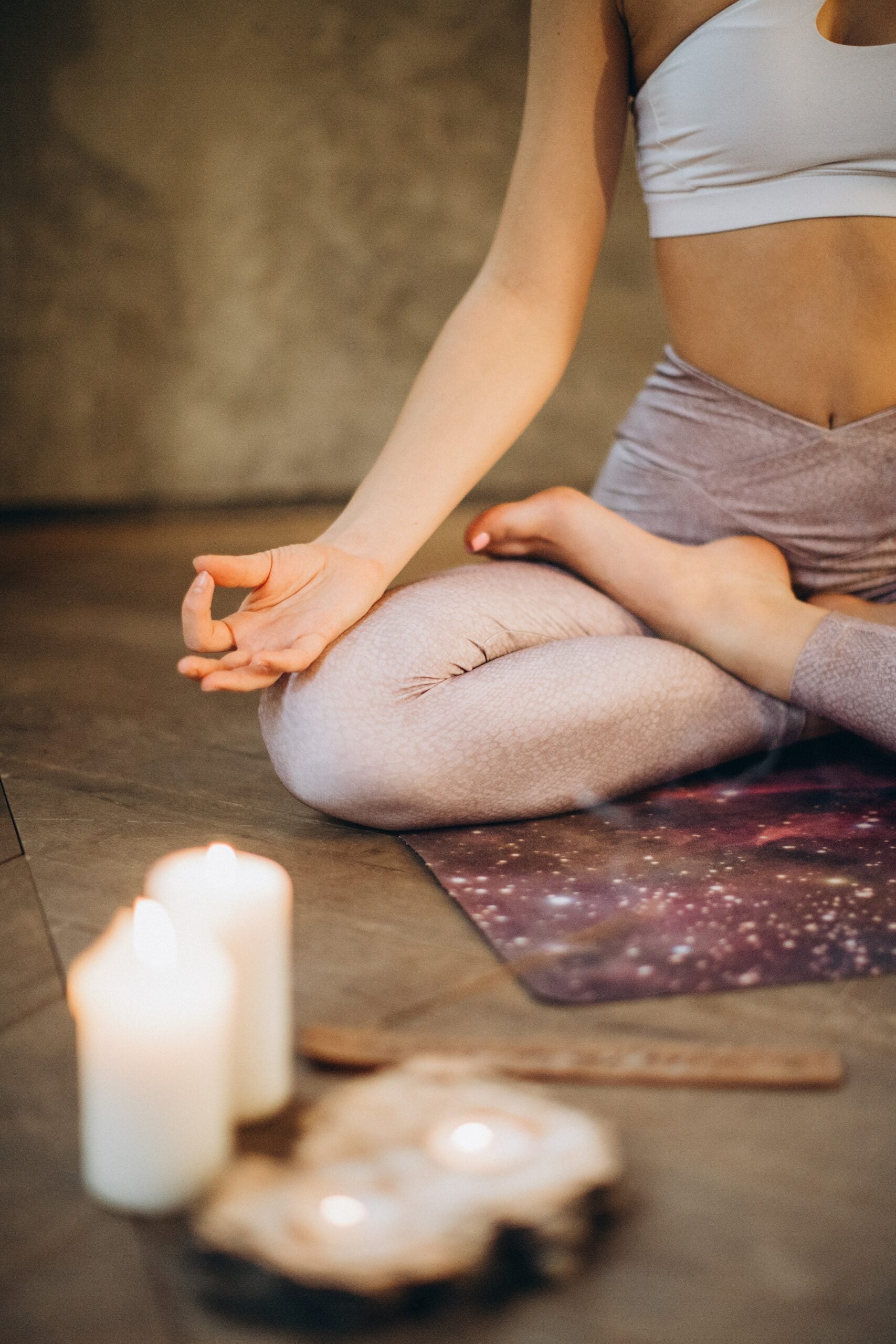 Person seated cross-legged in lotus pose meditating on a galaxy-pattern yoga mat with lit candles nearby in a calm indoor space