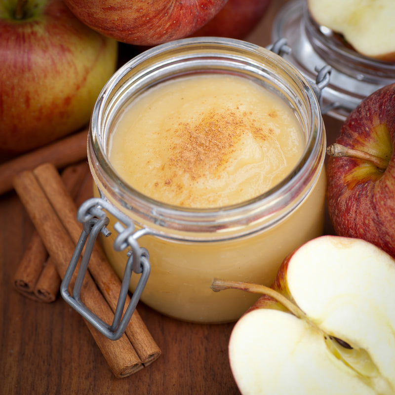 Jar of applesauce topped with cinnamon, surrounded by whole and halved red apples and cinnamon sticks on a wooden surface