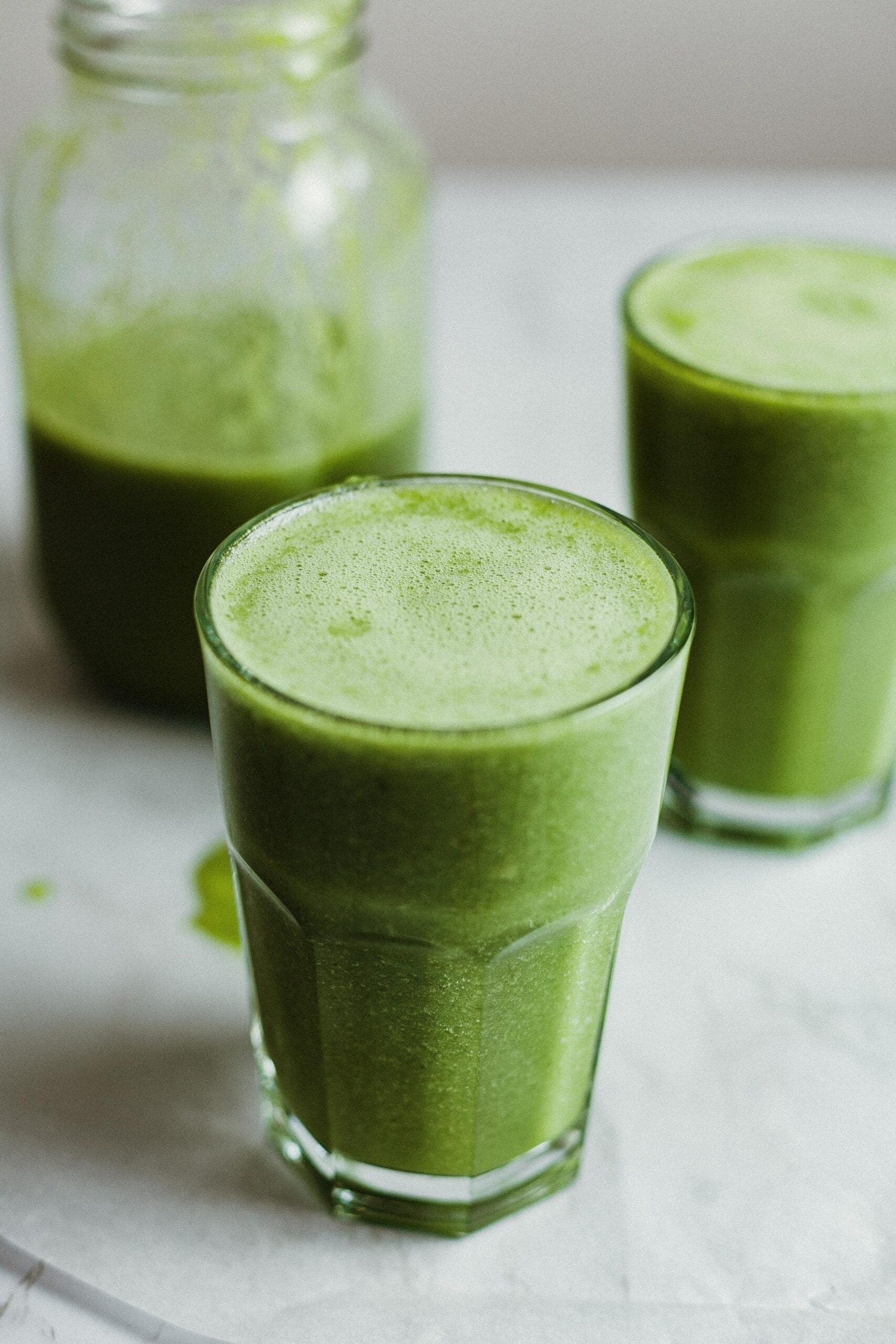 Close-up of two frothy green smoothies in glasses with a jar of green smoothie in the background on a light countertop