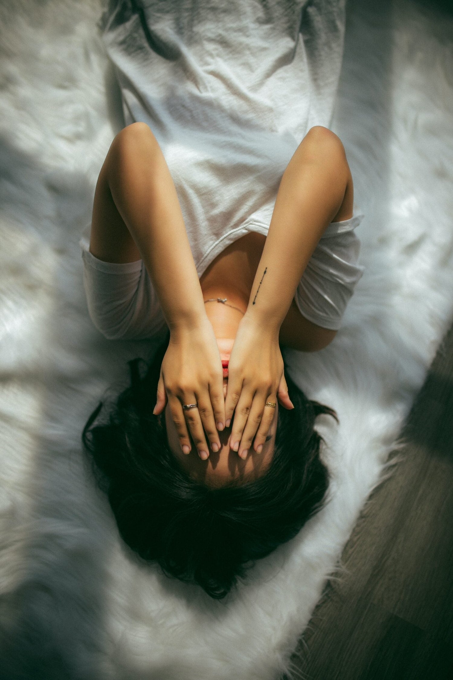 Woman lying on her back on a white rug covering her face with both hands, conveying headache or stress