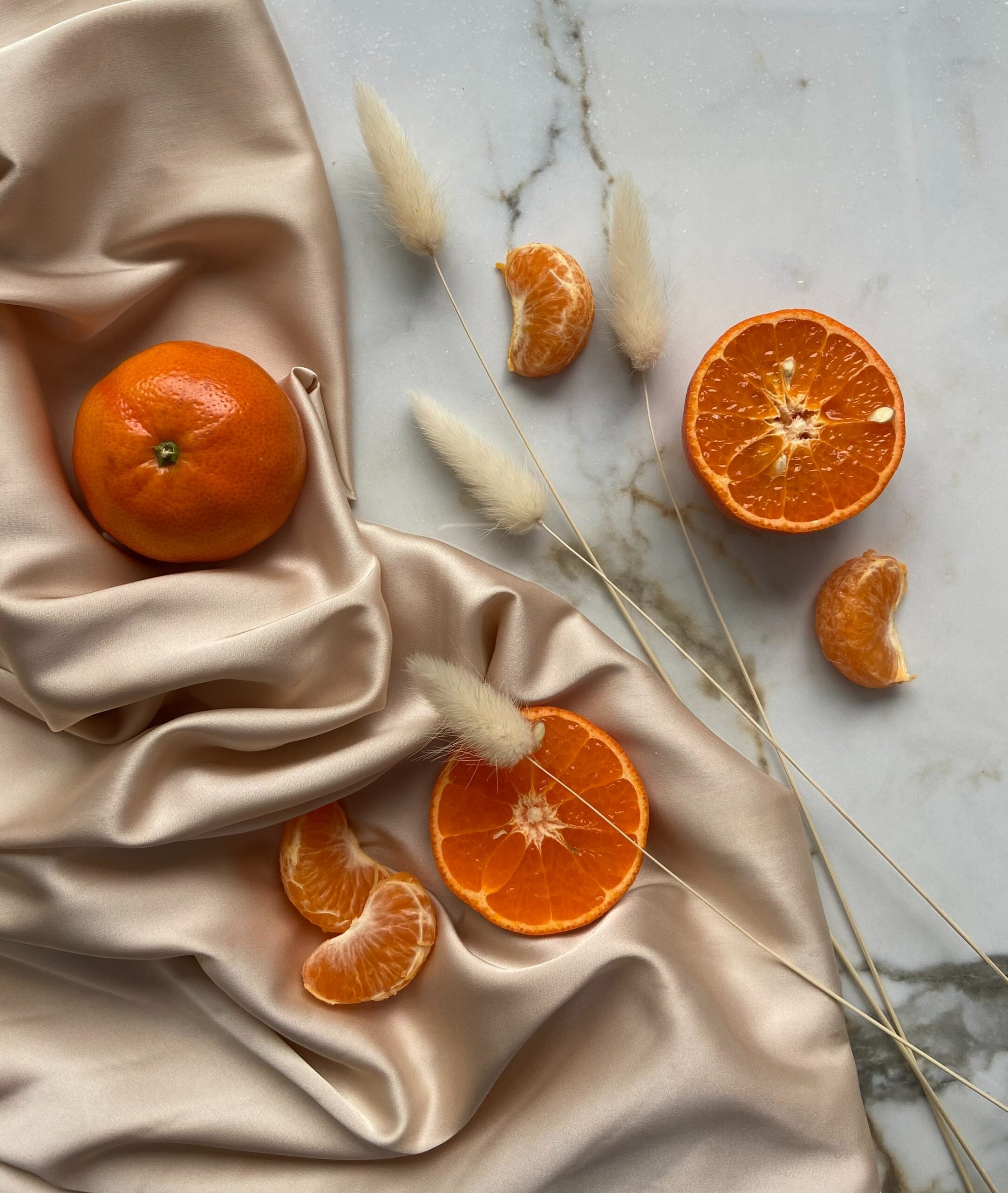 Top-down view of tangerines, a halved orange, and peeled segments arranged on pale satin and marble with dried bunny-tail grasses
