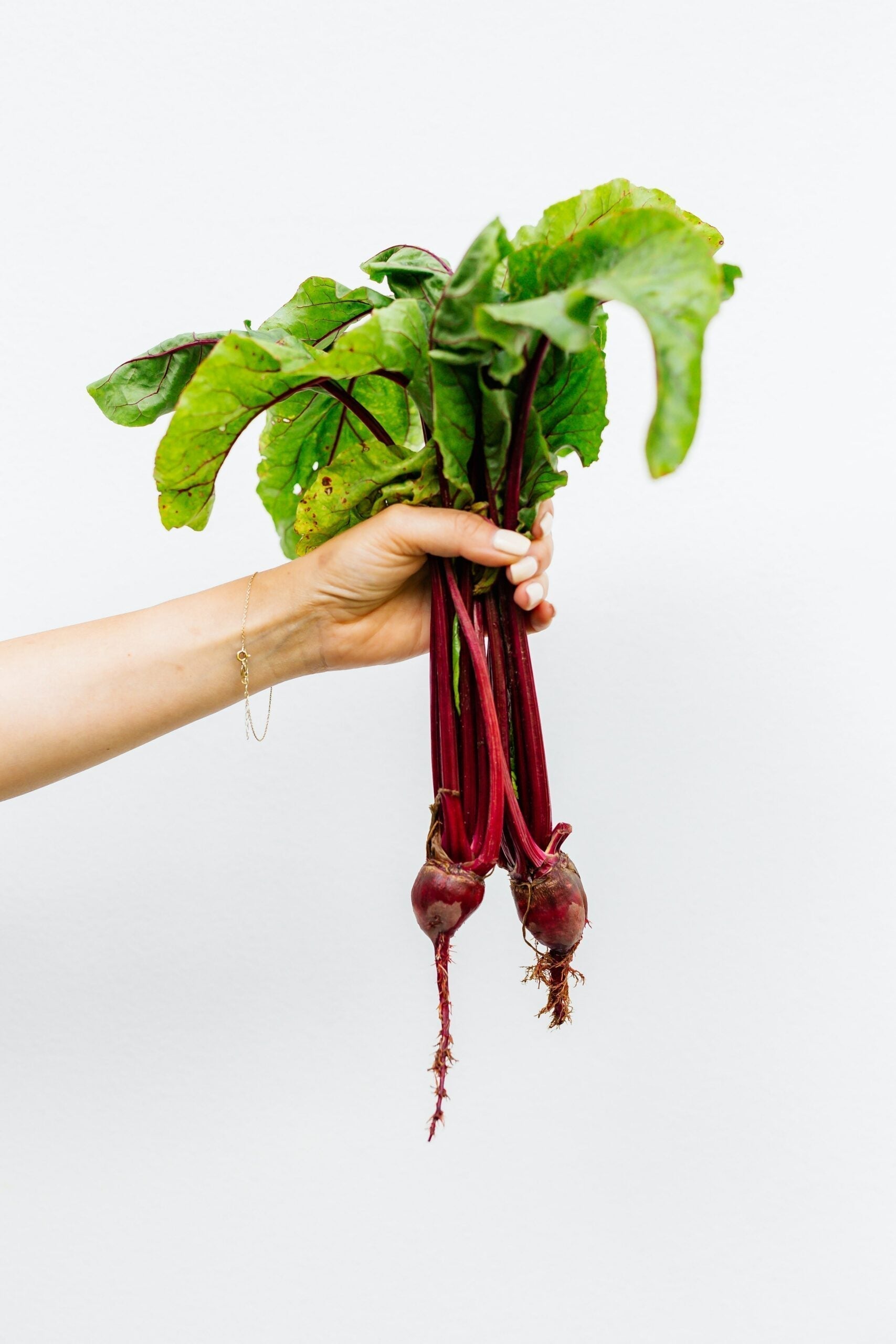 Hand holding two small beets with red stems and green leaves against a white background