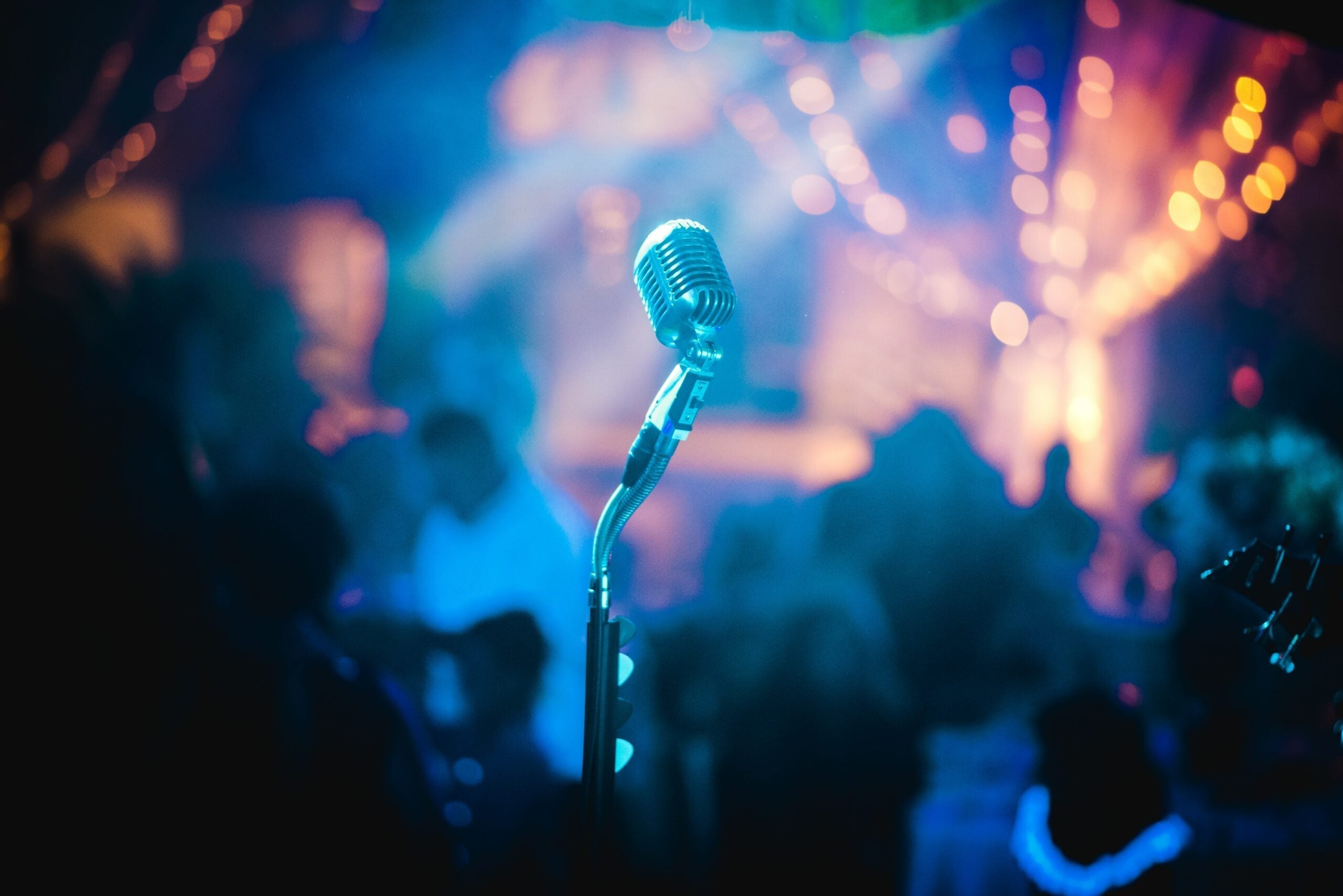 Vintage microphone on stand in foreground with blurred audience and warm string lights in a dim venue
