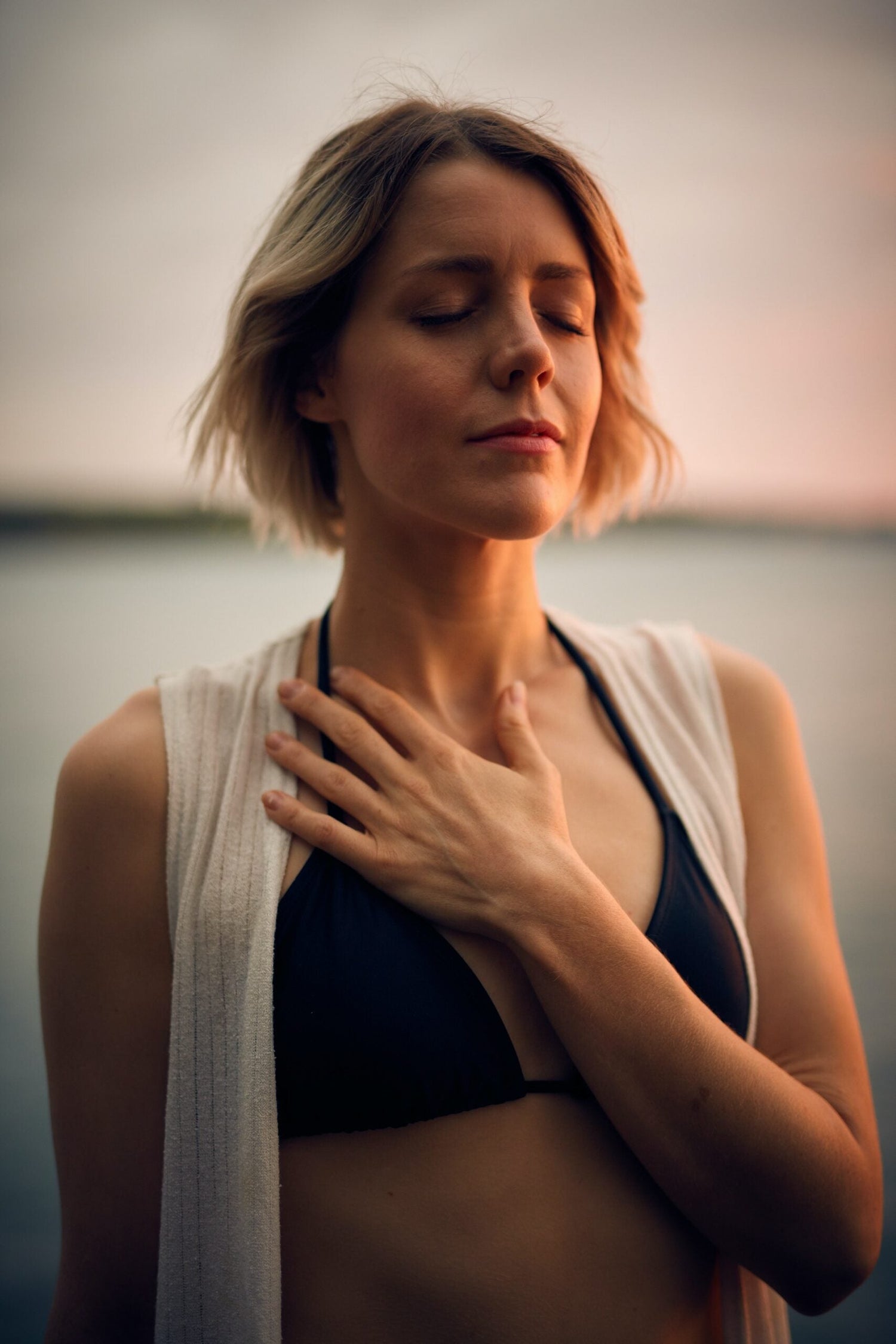 Woman with eyes closed placing a hand on her chest by calm water at sunset, appearing mindful and reflective