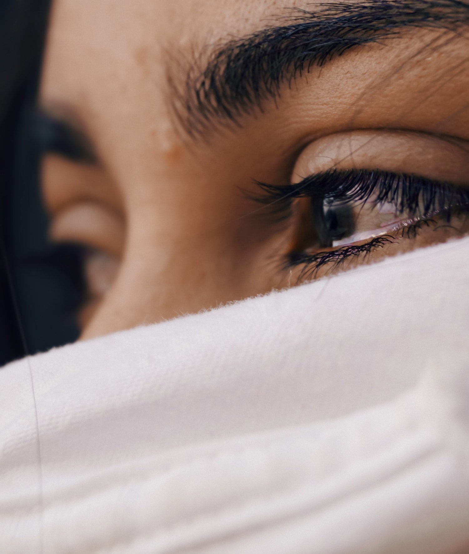 Close-up of a person’s eye looking sideways over a white cloth, showing eyelashes, eyebrow, and skin texture