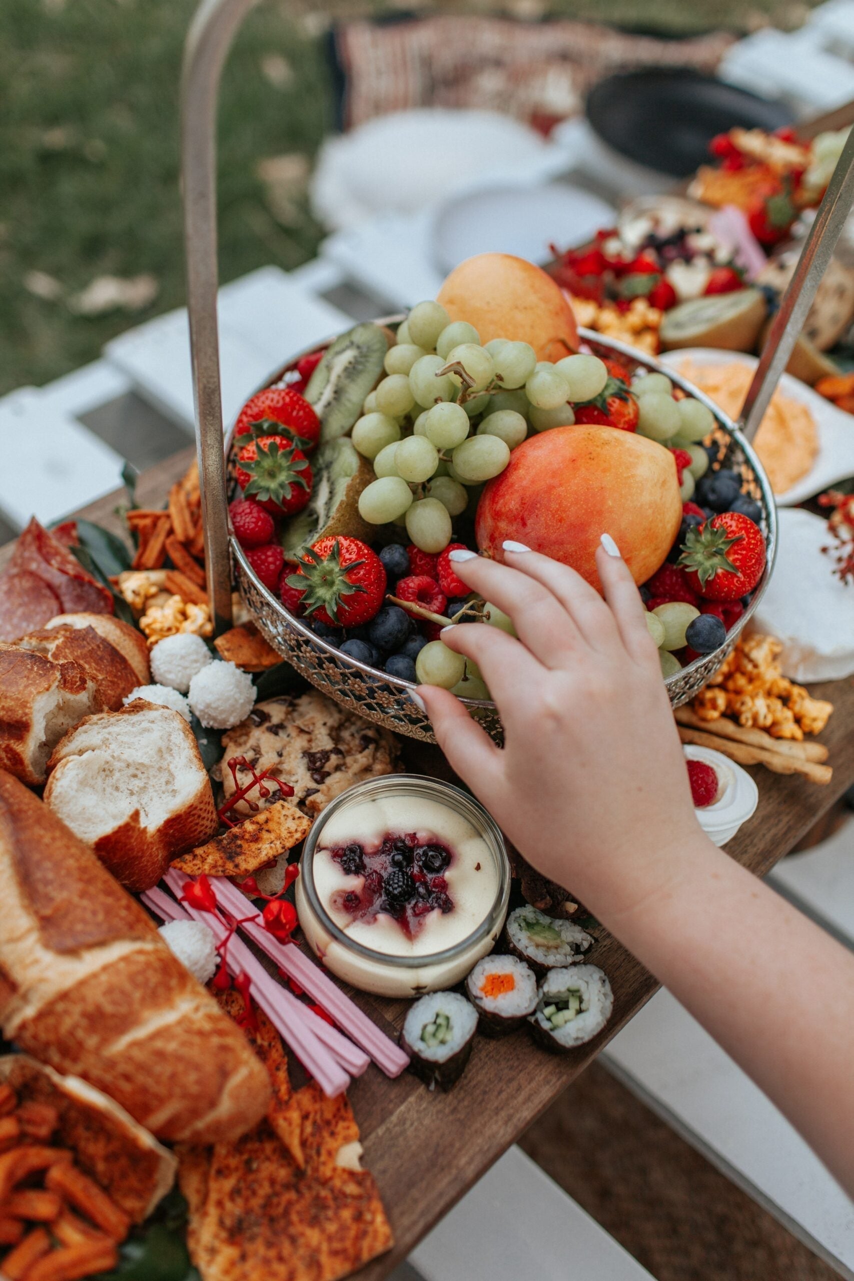 Hand reaching for grapes on a picnic-style breakfast board with fruit, pastries, sushi and yogurt outdoors