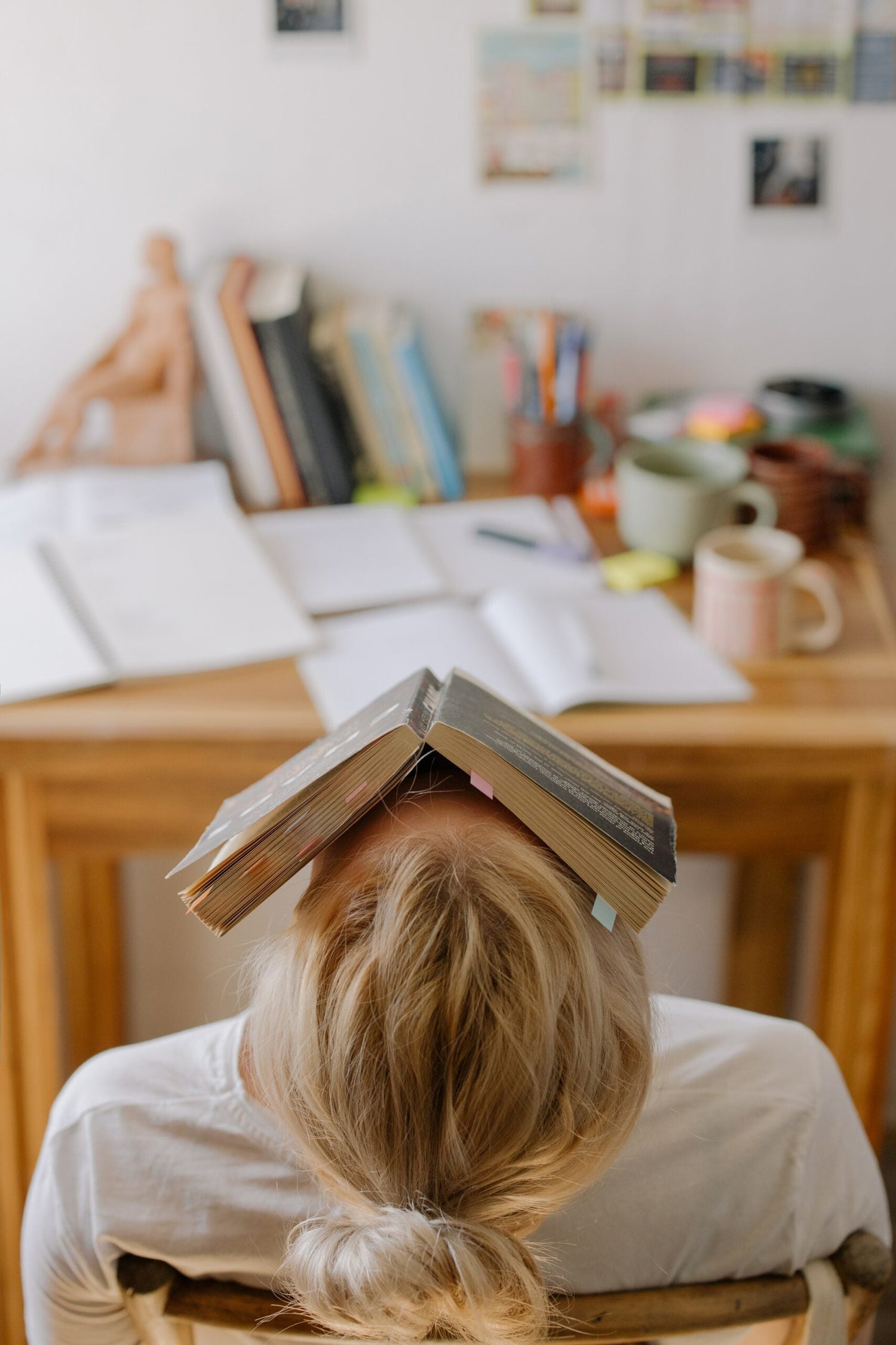 Person leaning back in a chair with an open book balanced on their forehead at a cluttered desk, suggesting study exhaustion