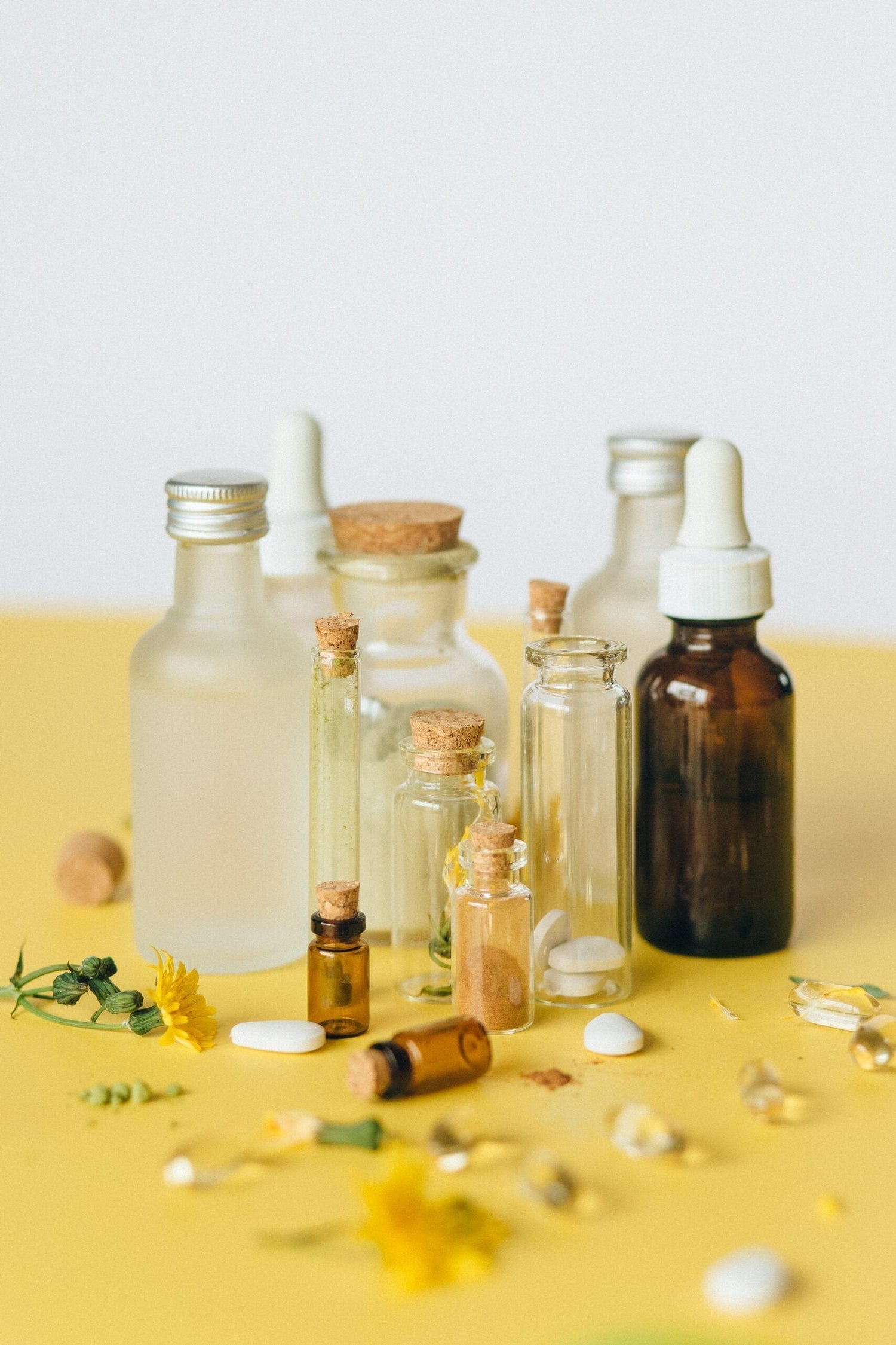 Assorted glass bottles and corked vials with droppers, dried herbs and yellow flowers arranged on a yellow tabletop