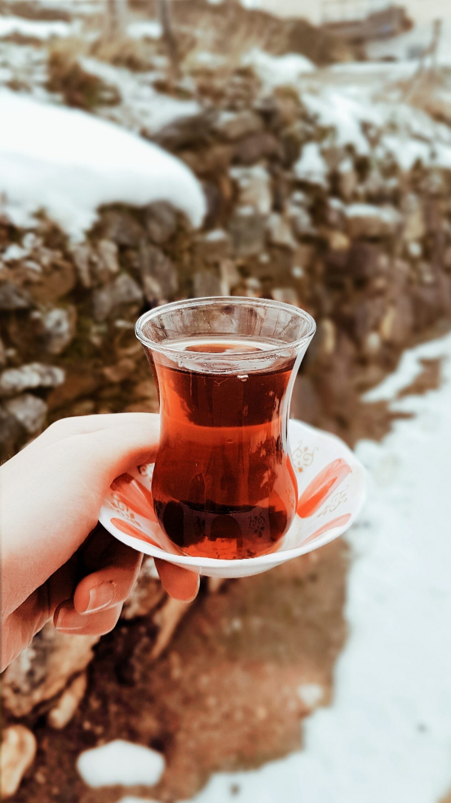 Hand holding a tulip-shaped glass of deep red hibiscus tea on a saucer outdoors with a snowy stone wall background