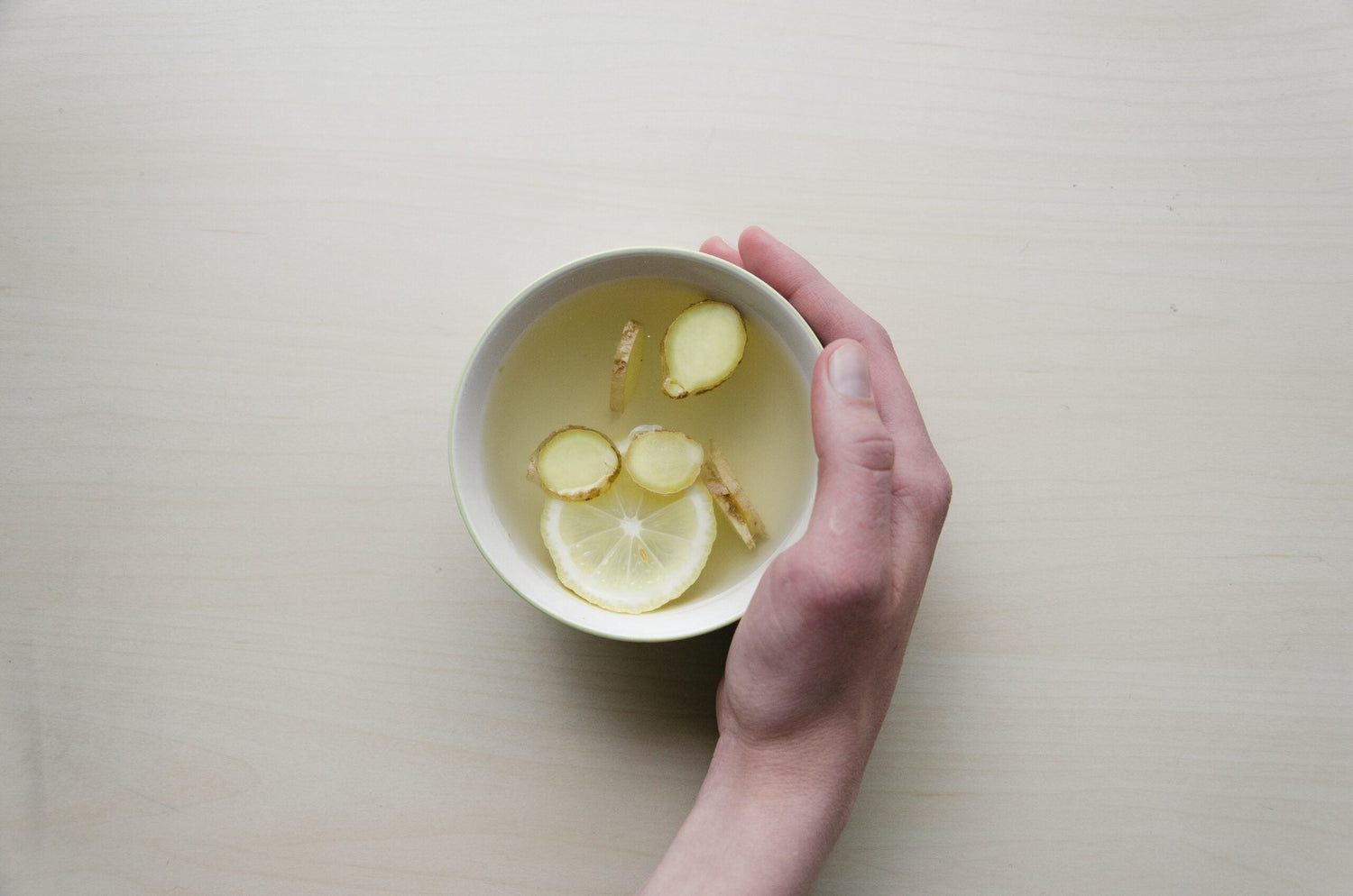 Hand holding a bowl of ginger tea with lemon slice and ginger pieces over a light wood table