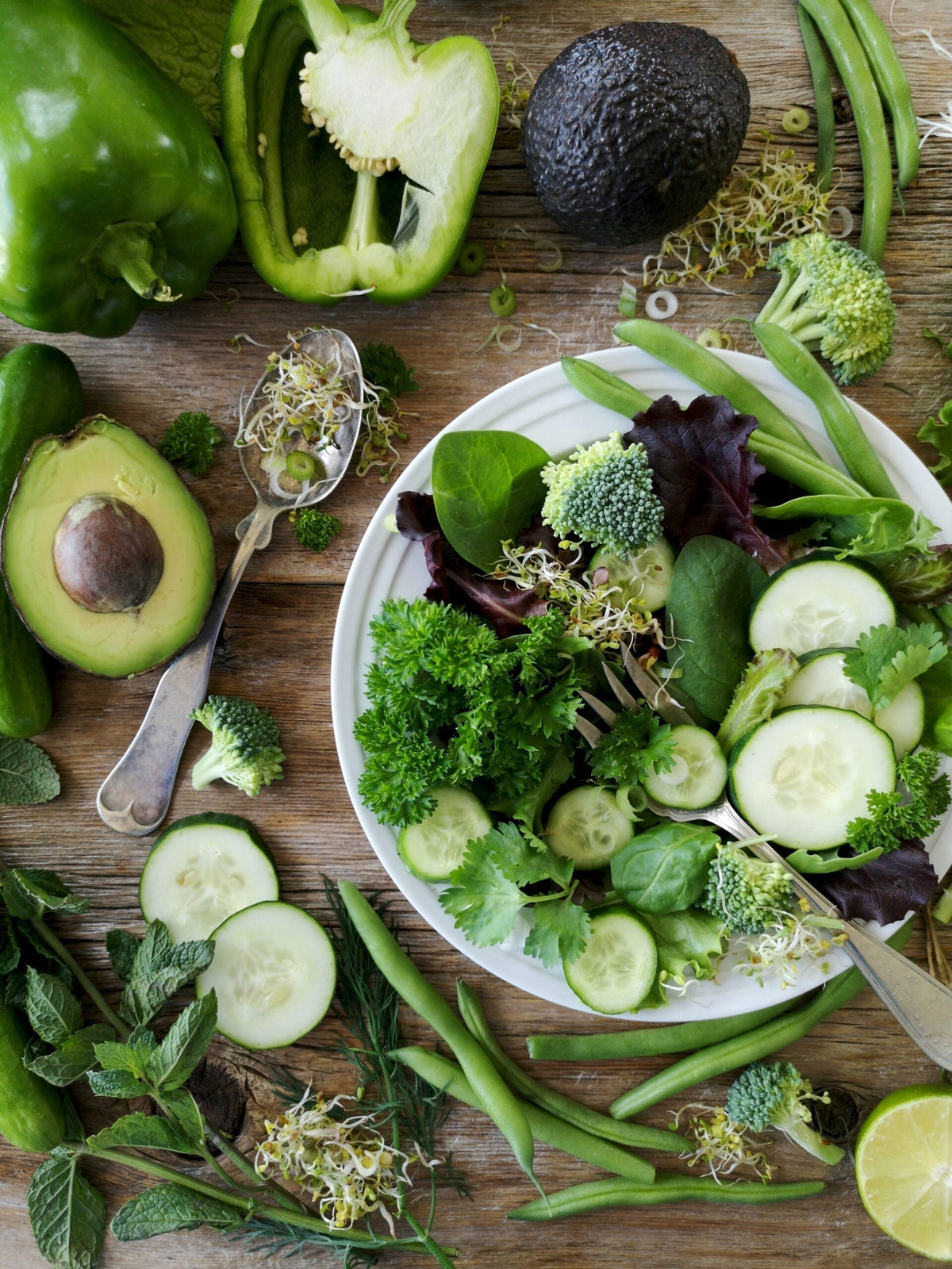 Bowl of mixed green salad with cucumber, avocado, broccoli, sprouts and herbs arranged on a wooden table