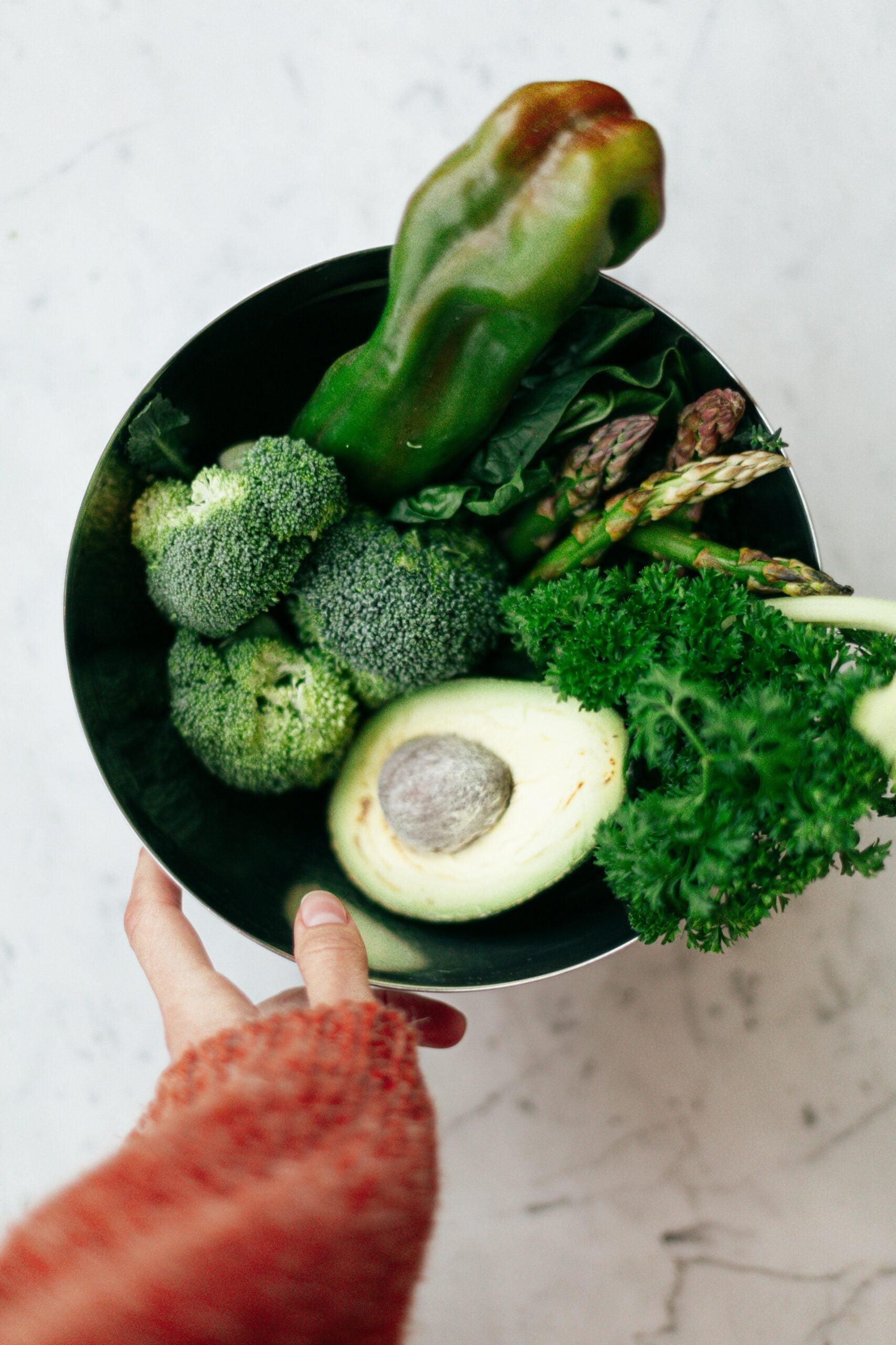 Hand holding a bowl of green vegetables including avocado, broccoli, asparagus, parsley, and a green pepper on a marble surface