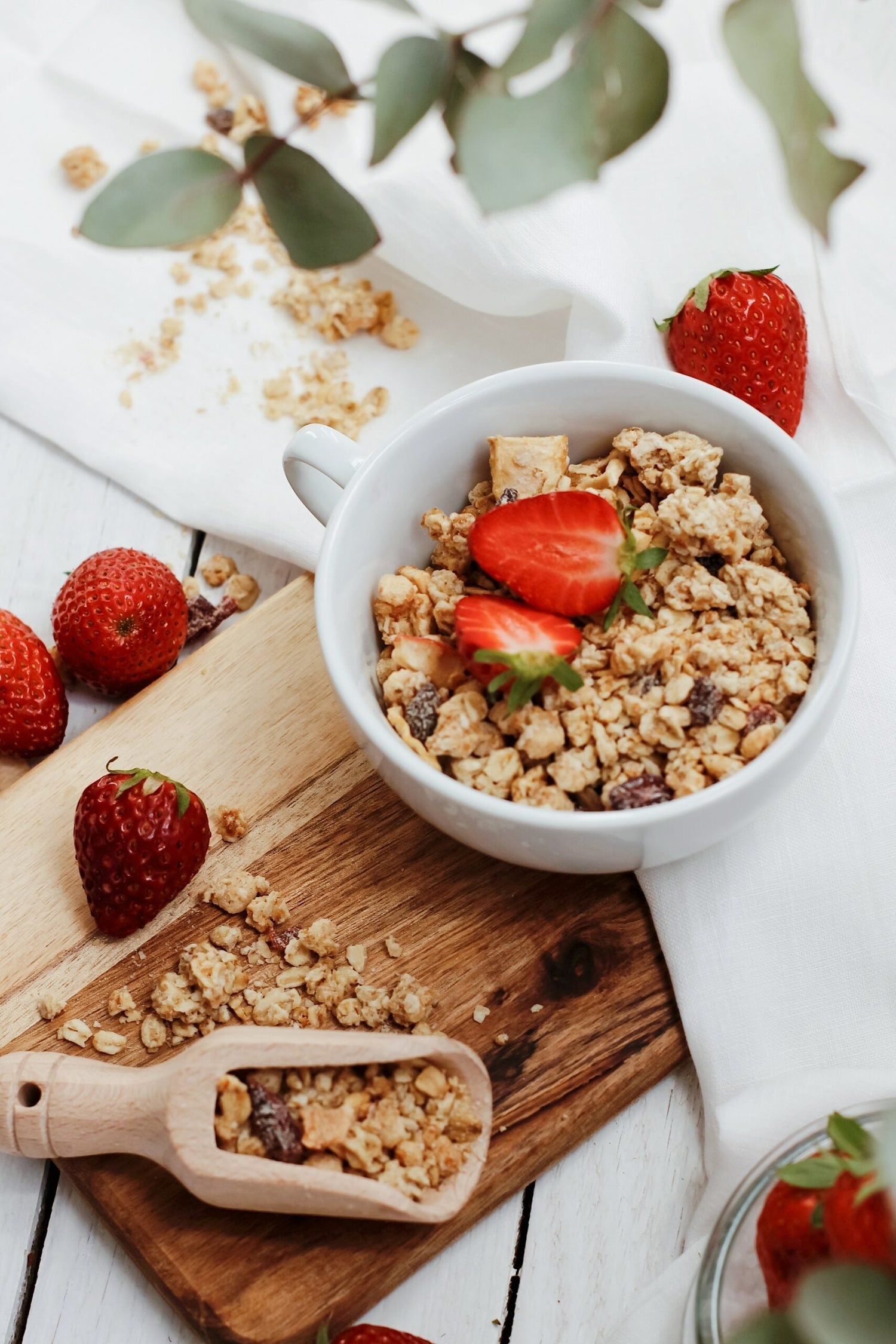 Bowl of granola topped with halved strawberries on a wooden board and white cloth, with scattered granola and whole strawberries nearby