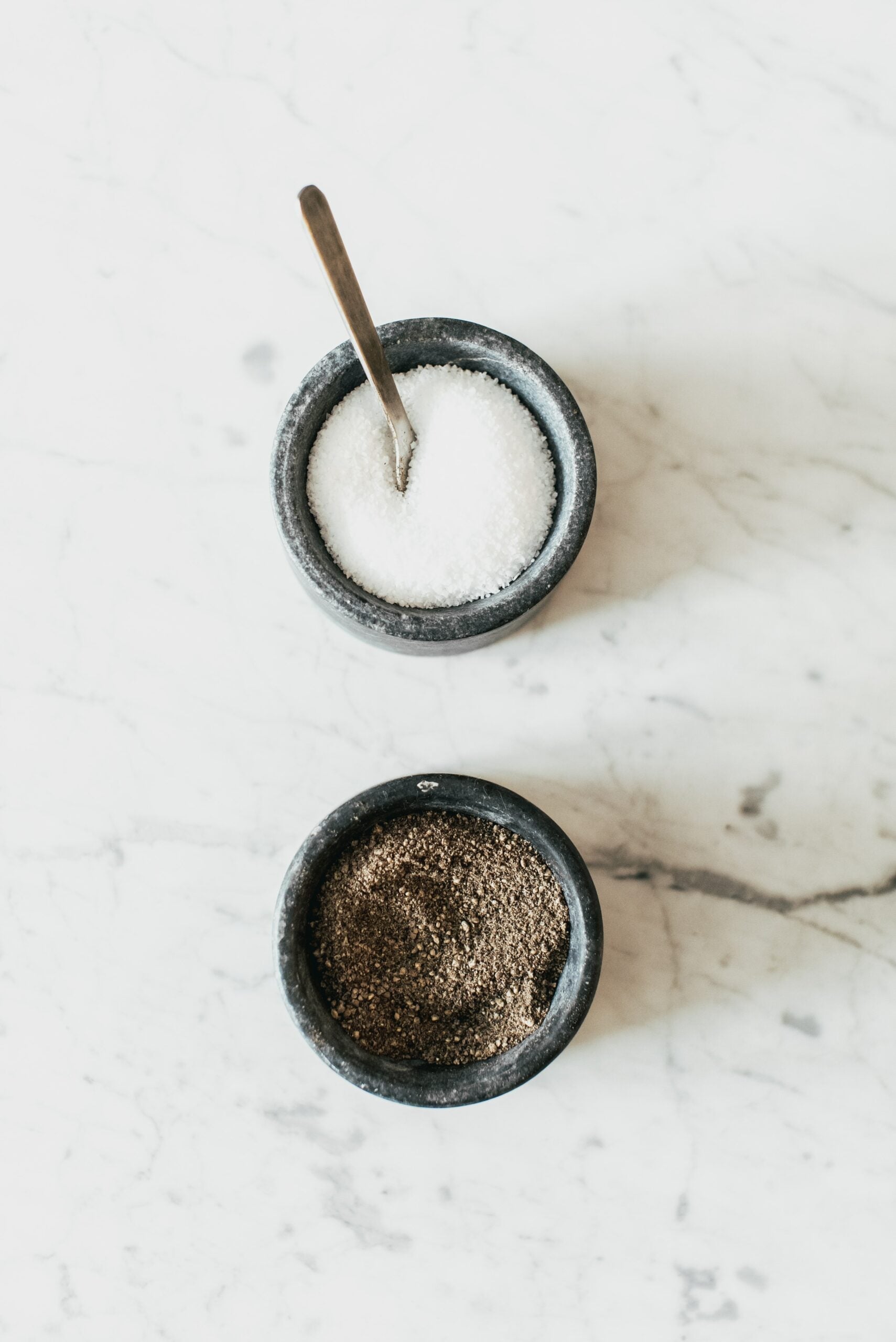 Two small stone bowls containing coarse salt and ground black pepper on a white marble countertop