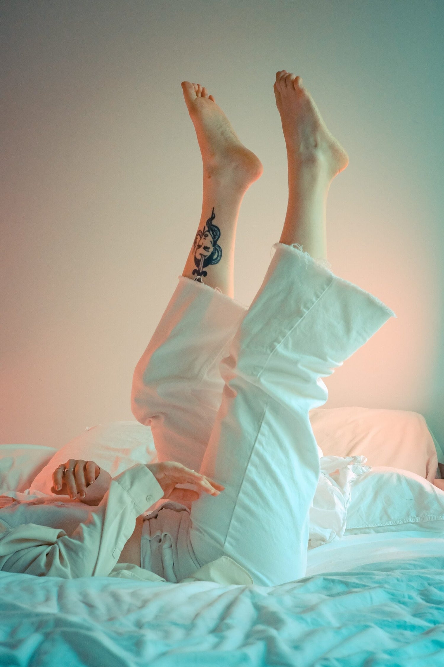 Person lying on bed with legs raised, wearing white pants and showing an ankle tattoo, soft teal and pink bedside lighting