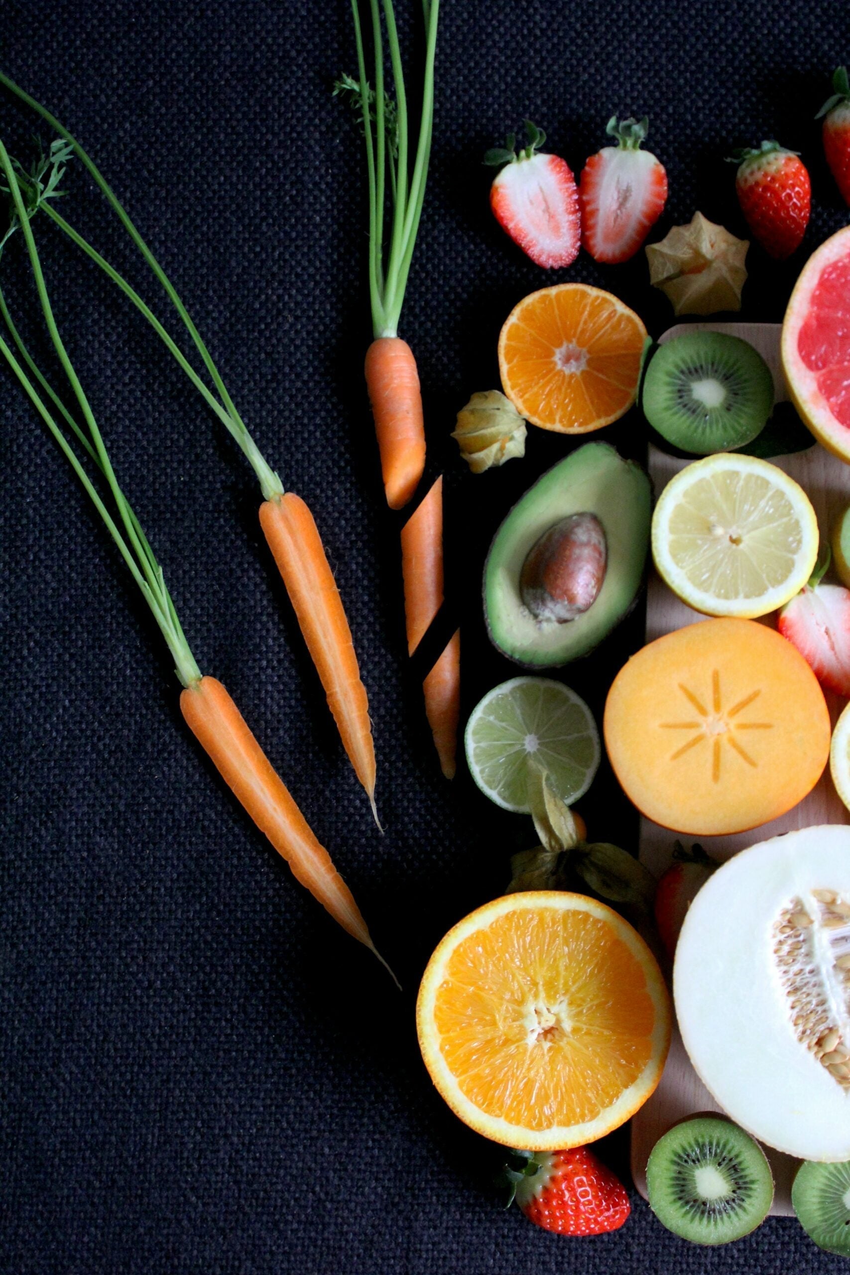 Assorted fiber-rich fruits and vegetables on a dark cloth, including baby carrots, halved avocado, orange, kiwi, strawberries, and melon