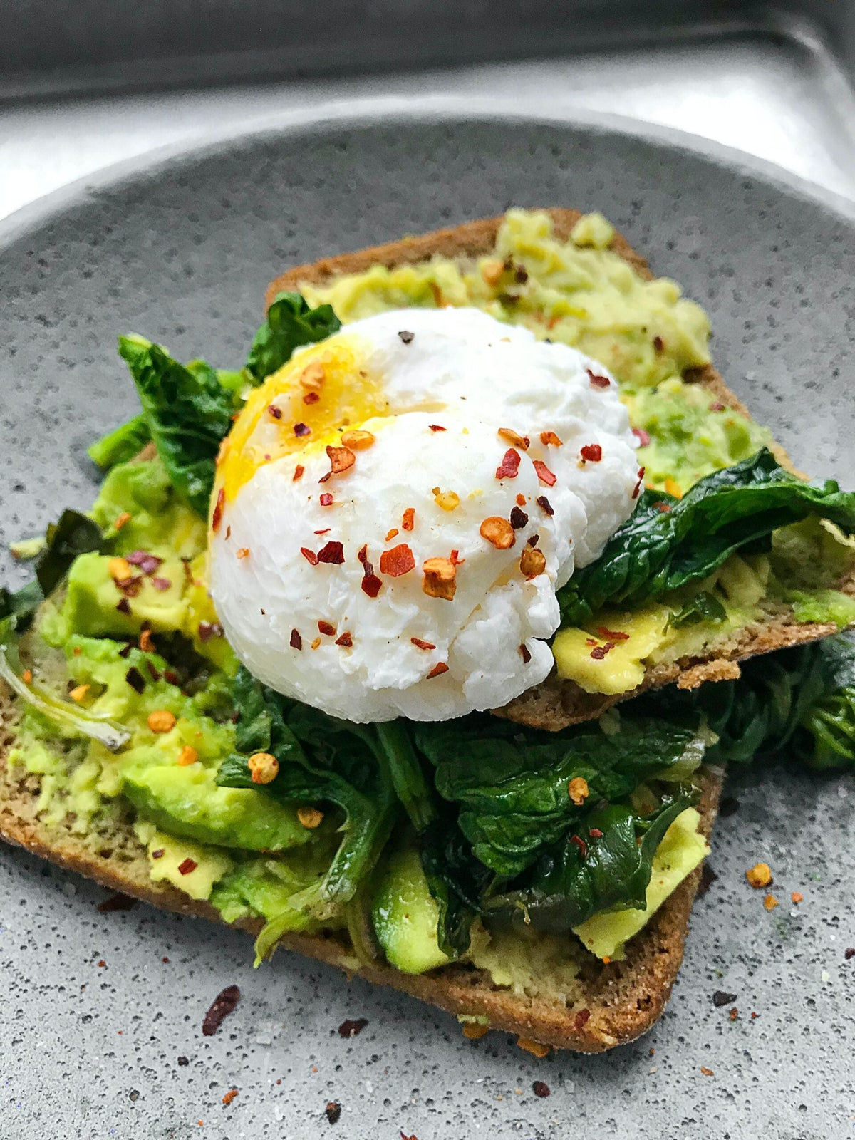 Close-up of avocado toast with a poached egg, spinach, and chili flakes on a gray plate