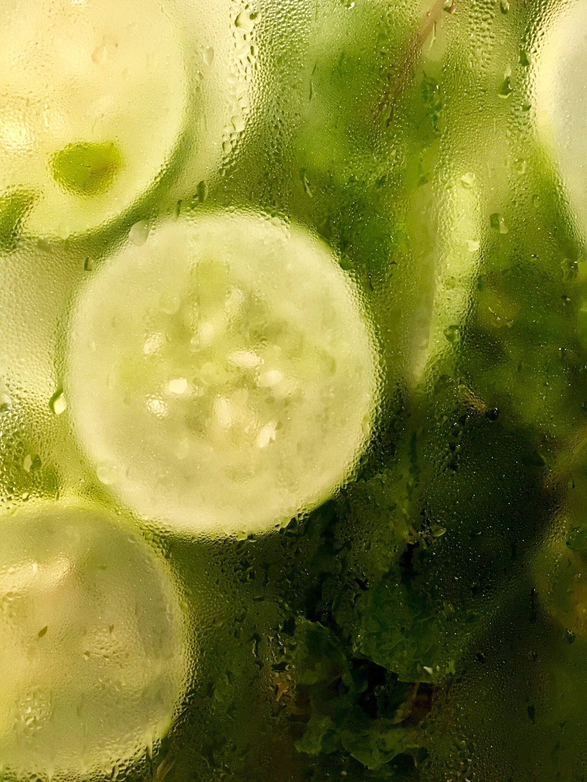 Close-up of cucumber slices and green herbs steeping in water behind condensation-covered glass