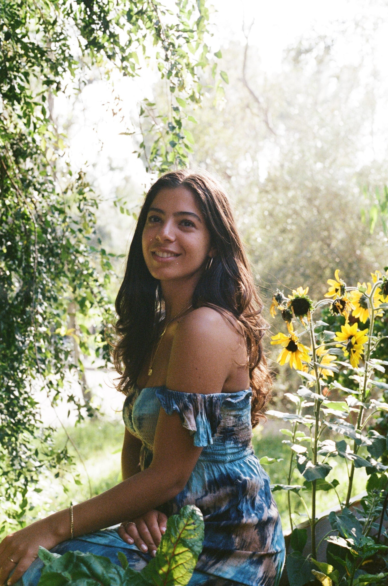 Young woman smiling while sitting in a sunlit garden beside yellow sunflowers, wearing a blue off-shoulder dress